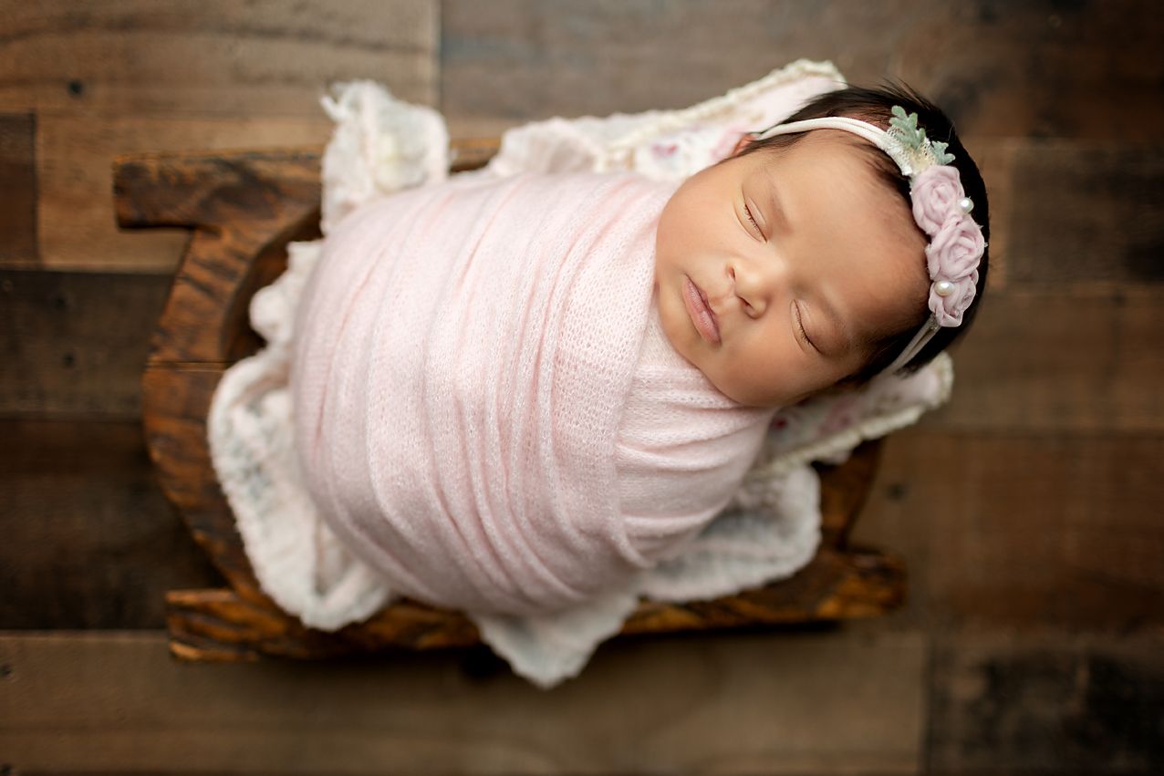 Newborn baby wrapped in pink blanket, asleep in a wooden bowl, floral headband.