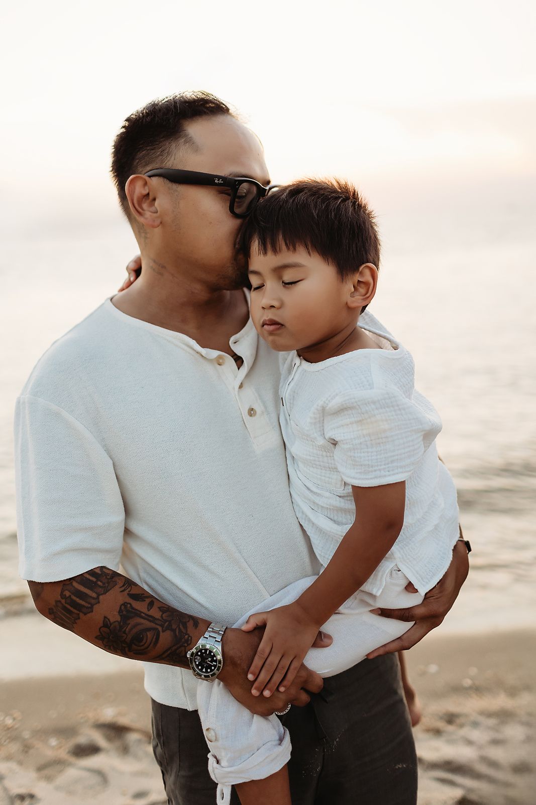 Father holding a sleeping child, beach setting. Man wearing sunglasses, white shirt; child in white. Warm light.