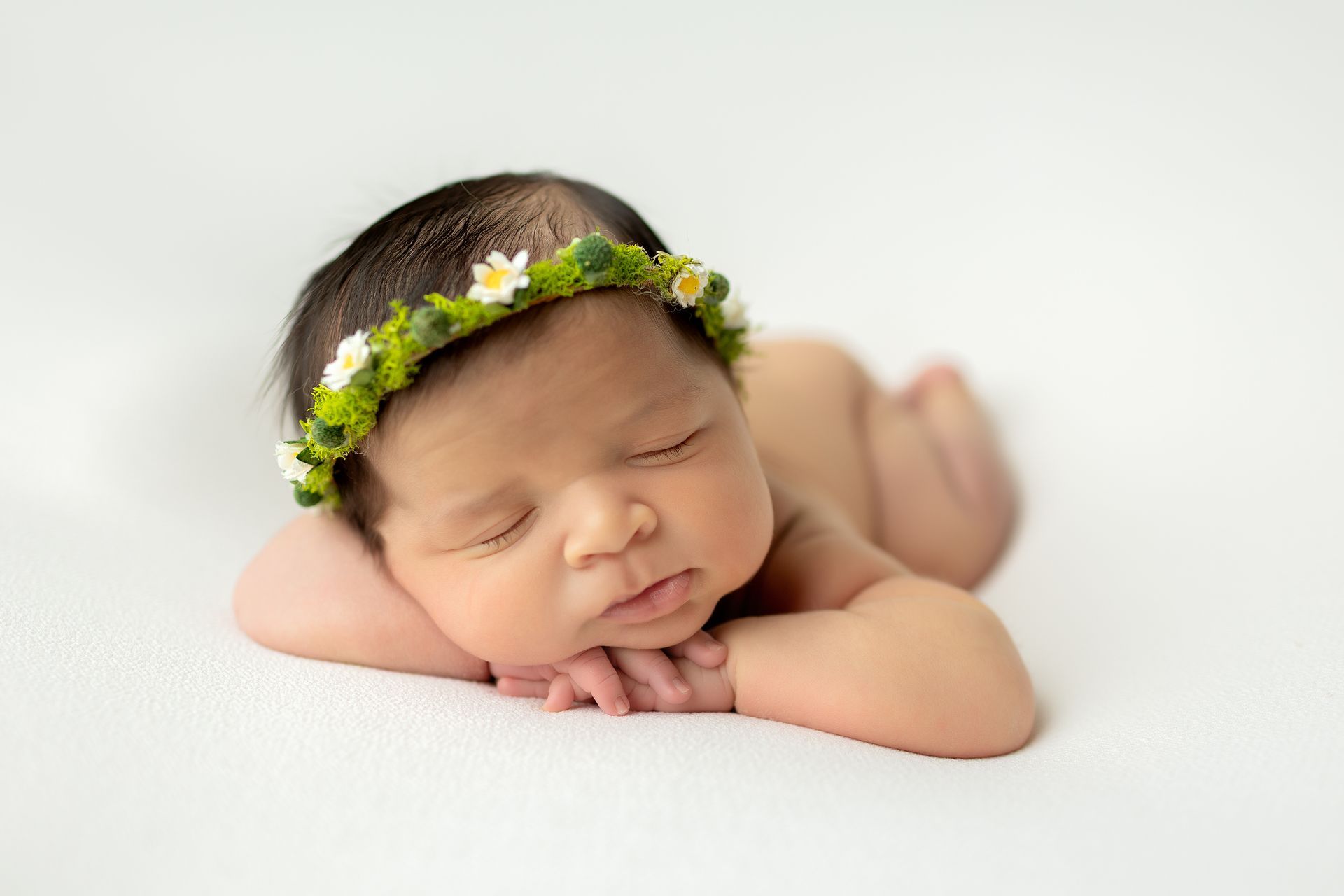 Newborn wearing a flower crown, sleeping on a white surface.