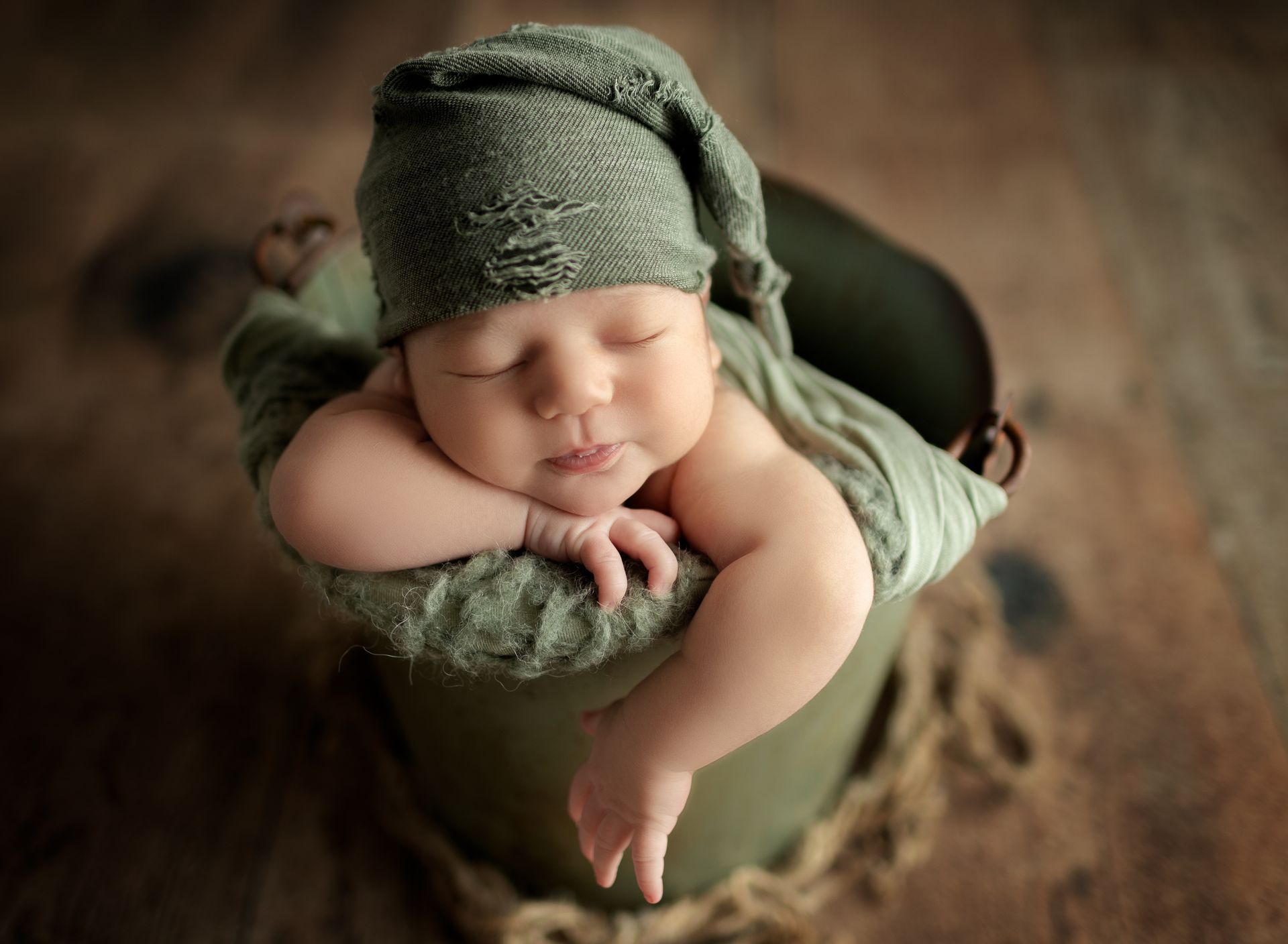 Sleeping baby wearing green cap and swaddled in green fabric, nestled in a metal bucket on a wood surface.