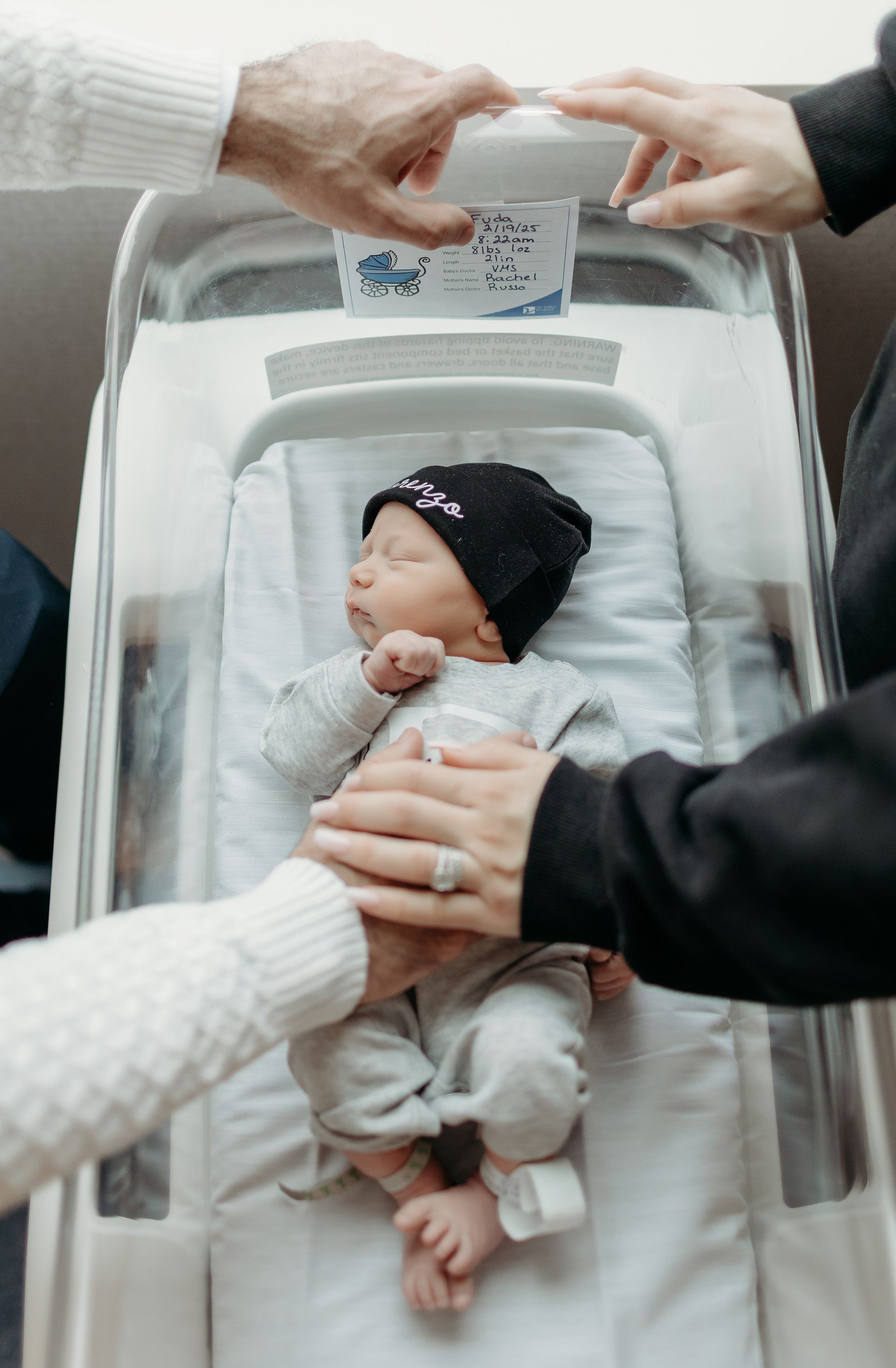 Newborn baby in a hospital bassinet, surrounded by hands. Baby wearing a black hat and grey onesie.