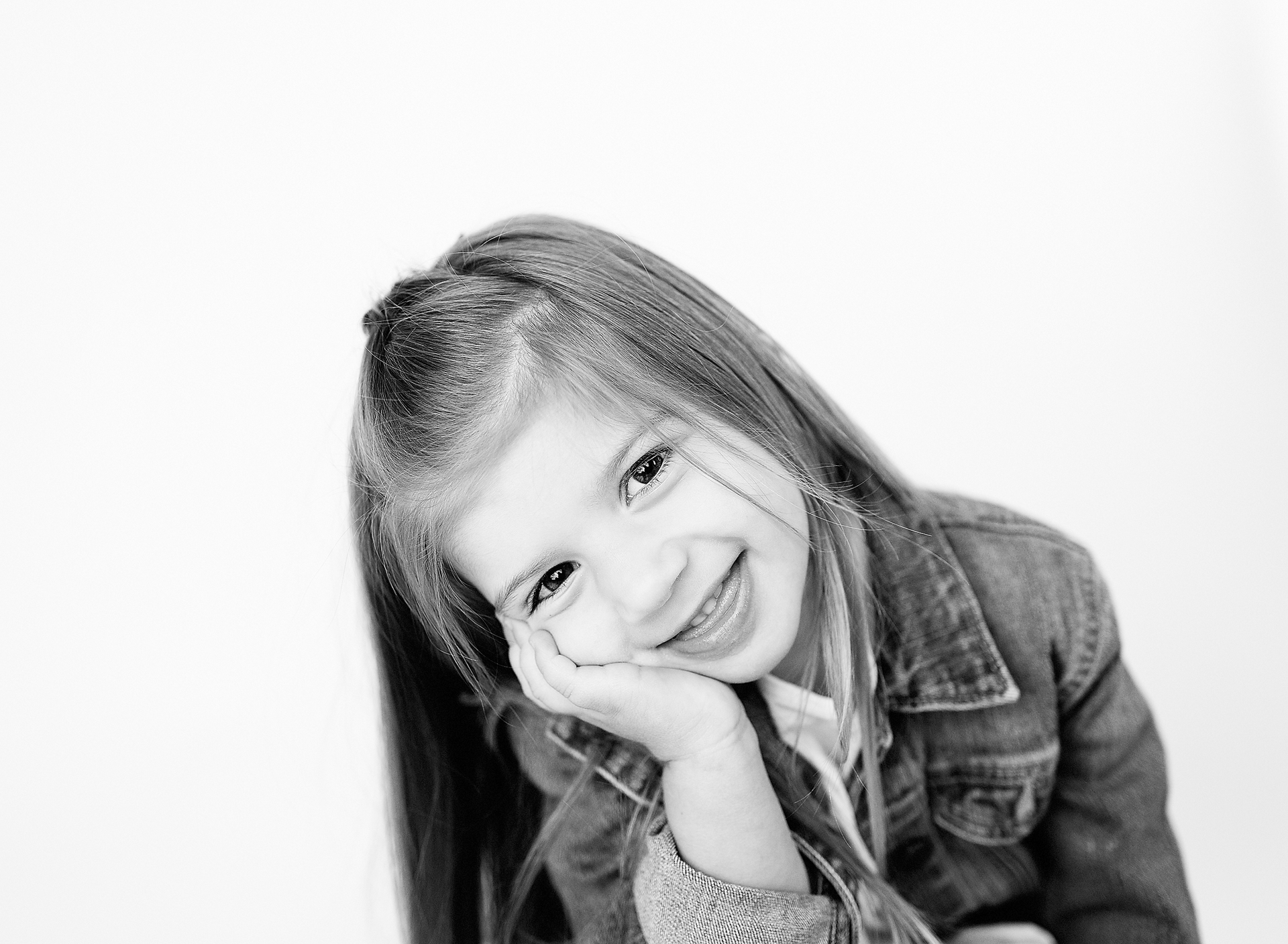 Girl with long hair, smiling, resting her chin on her hand, wearing a denim jacket, against a white background.
