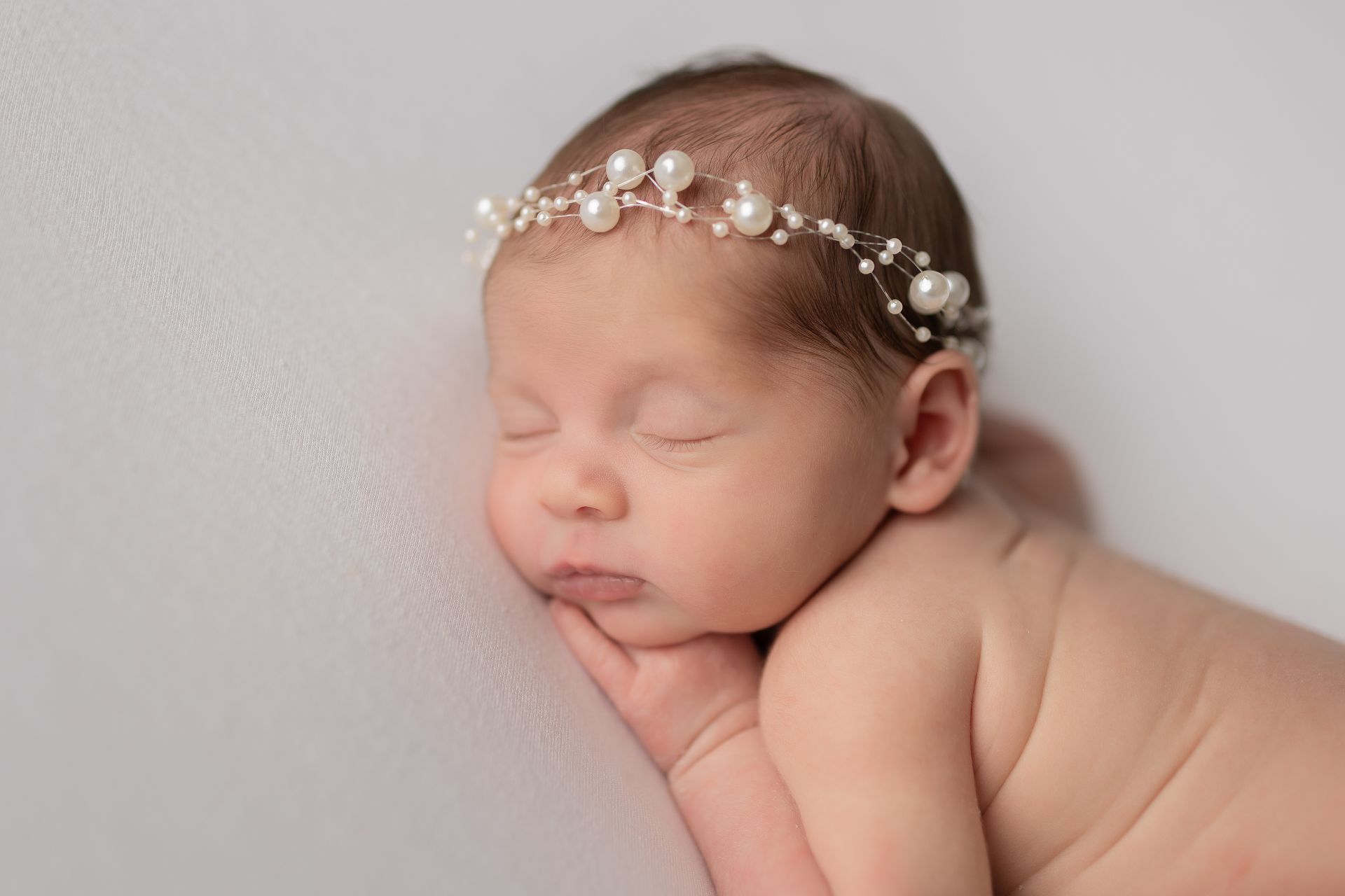 A newborn baby wearing a pearl headband is sleeping on a white blanket.