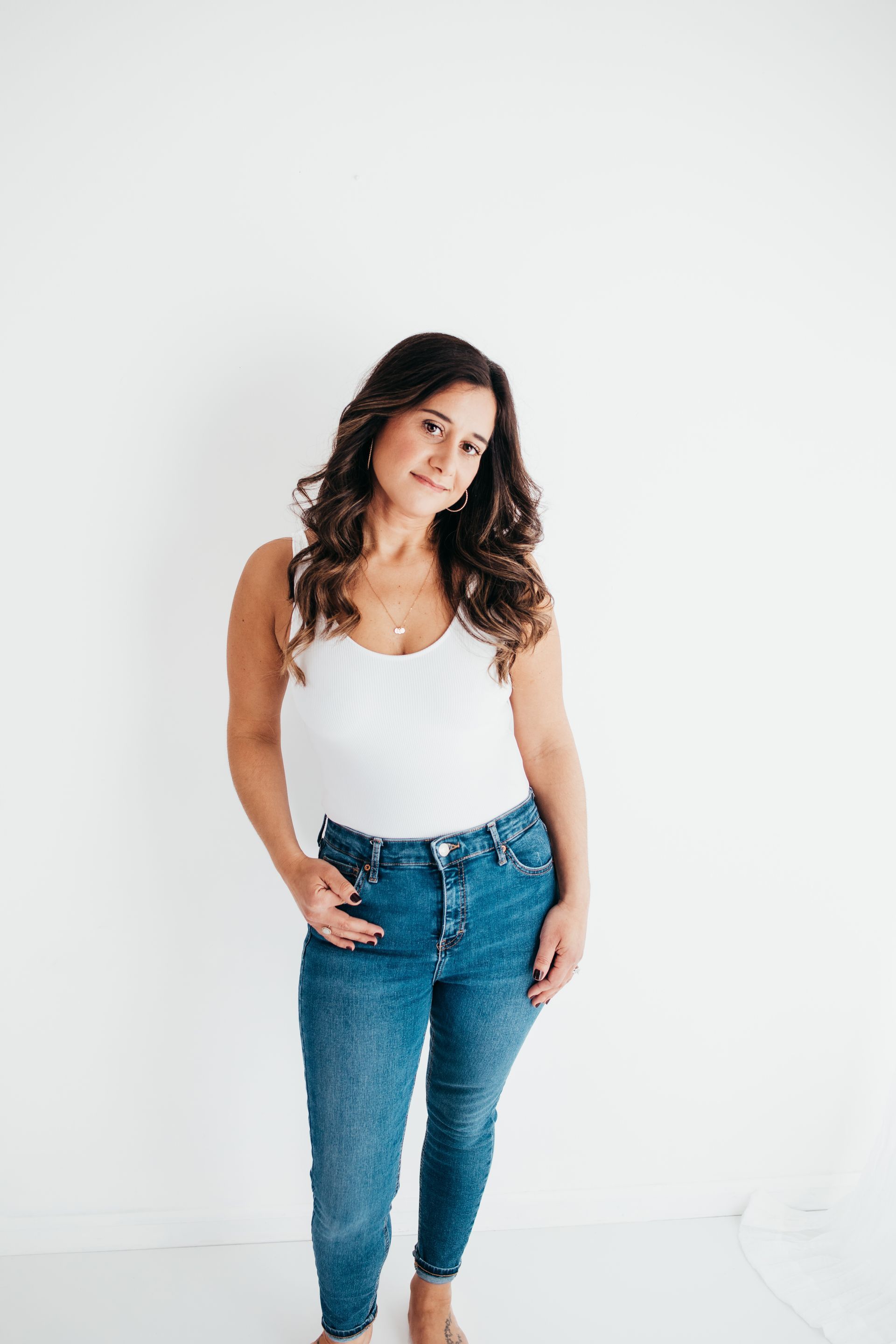 Lifestyle photographer capturing a woman in a white tank top and blue jeans standing in front of a white wall.