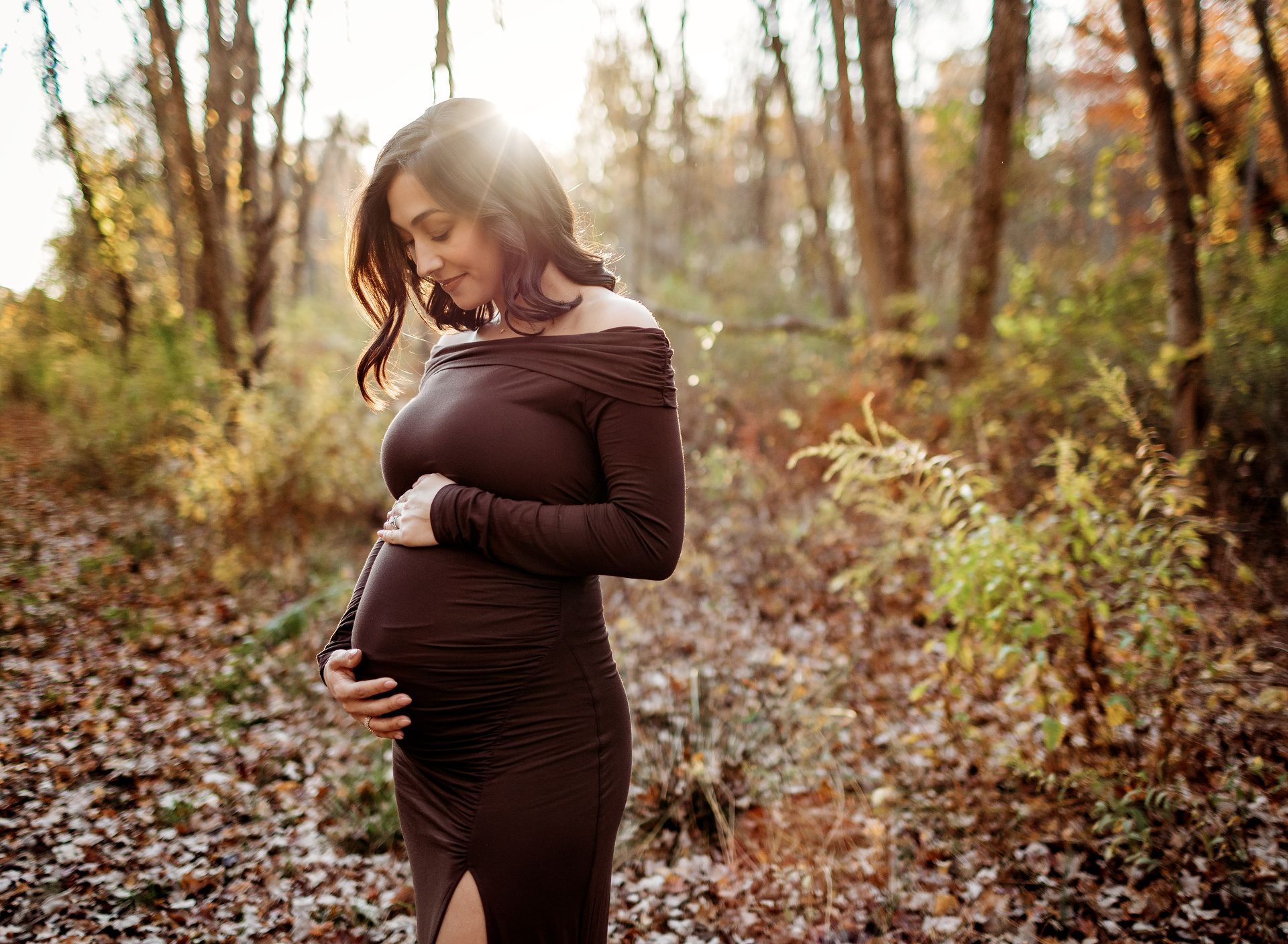 Pregnant person in a brown dress, standing in a sunlit forest, holding their belly.