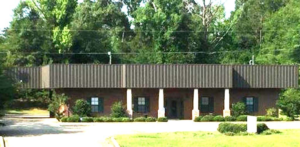 A low, brick building with a dark facade, white columns, and green shutters, set in front of trees.