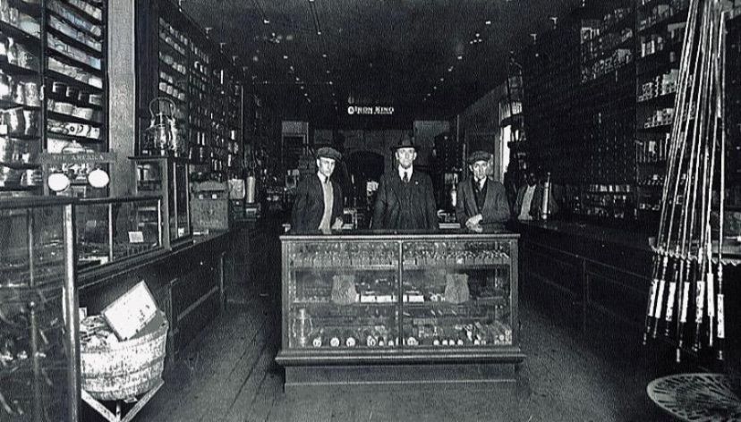 Inside an old shop with shelves of merchandise, three men stand behind a display case, looking at the camera.