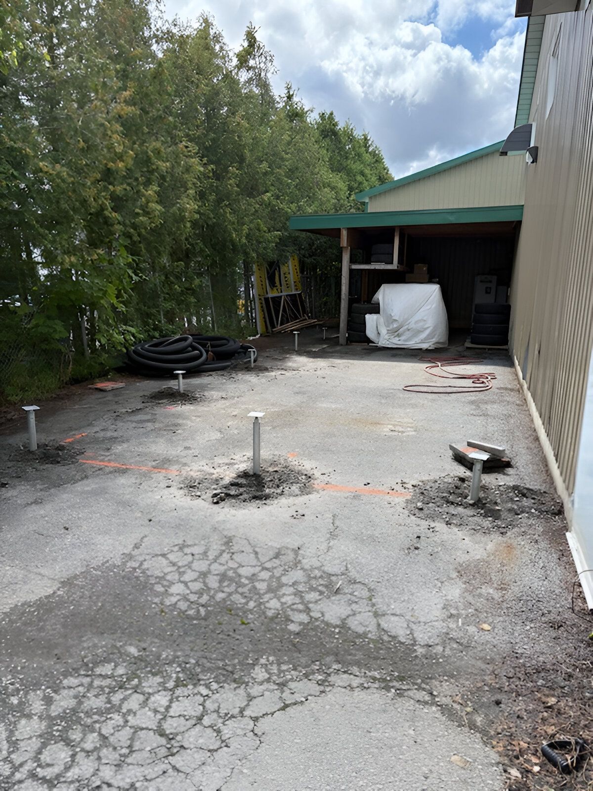 Allée fissurée à côté d'un garage, avec une petite souche d'arbre et des pneus empilés sous un abri de voiture au toit vert.
