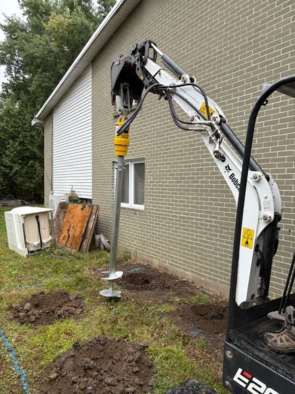 Une excavatrice effectue des forages à côté d'une maison en briques, avec des tas de terre et une tarière jaune au niveau des fondations.