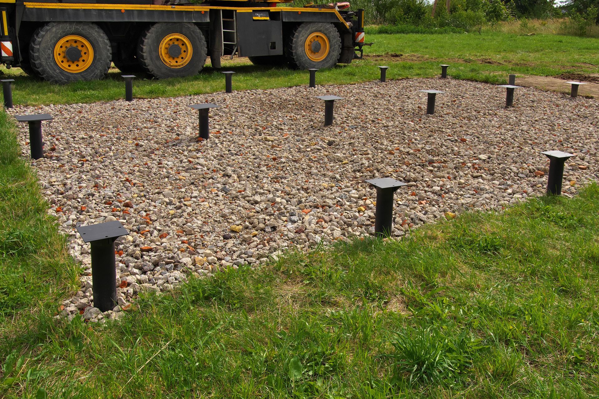 Fondation en gravier avec poteaux de soutien noirs, installée dans l'herbe à côté d'un ensemble de roues de remorque stationné.