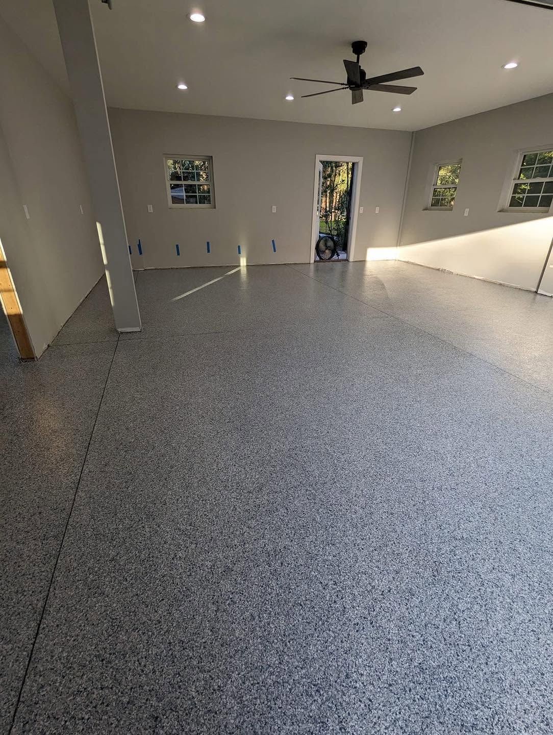 Empty modern room with gray speckled floor, ceiling fan, windows, and open door to outside.