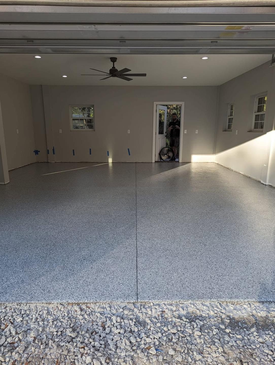 Empty garage with speckled epoxy floor, ceiling fan, and open doorway to a leafy yard.
