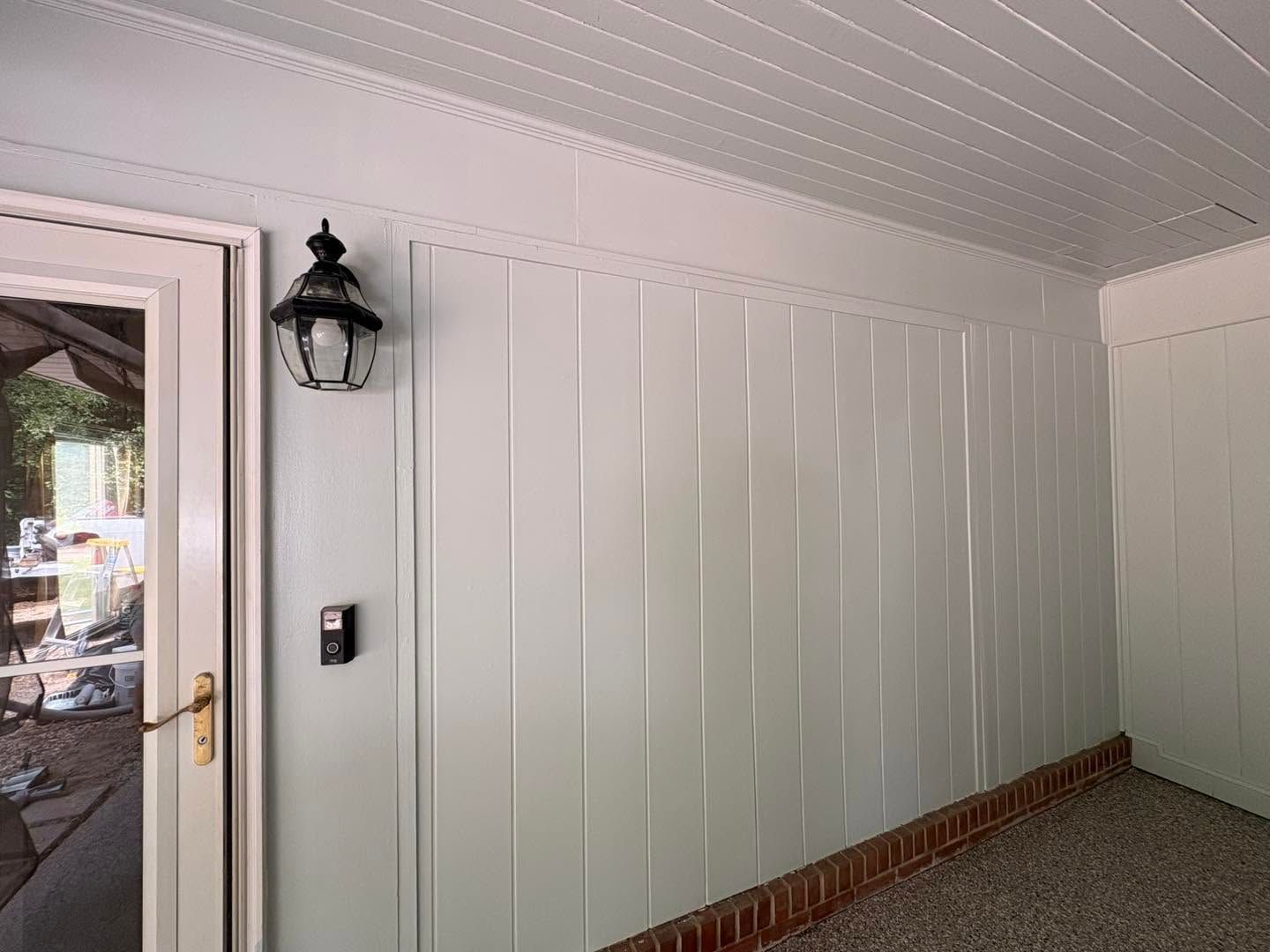 Front porch with white paneled wall, black lantern, and glass door at left; brick trim along the floor.