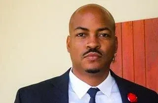 Portrait of a man in a dark suit and tie with a red lapel pin, standing before a wooden wall