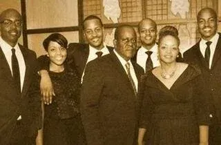 Group of six people posing indoors in formal black attire, smiling at the camera.