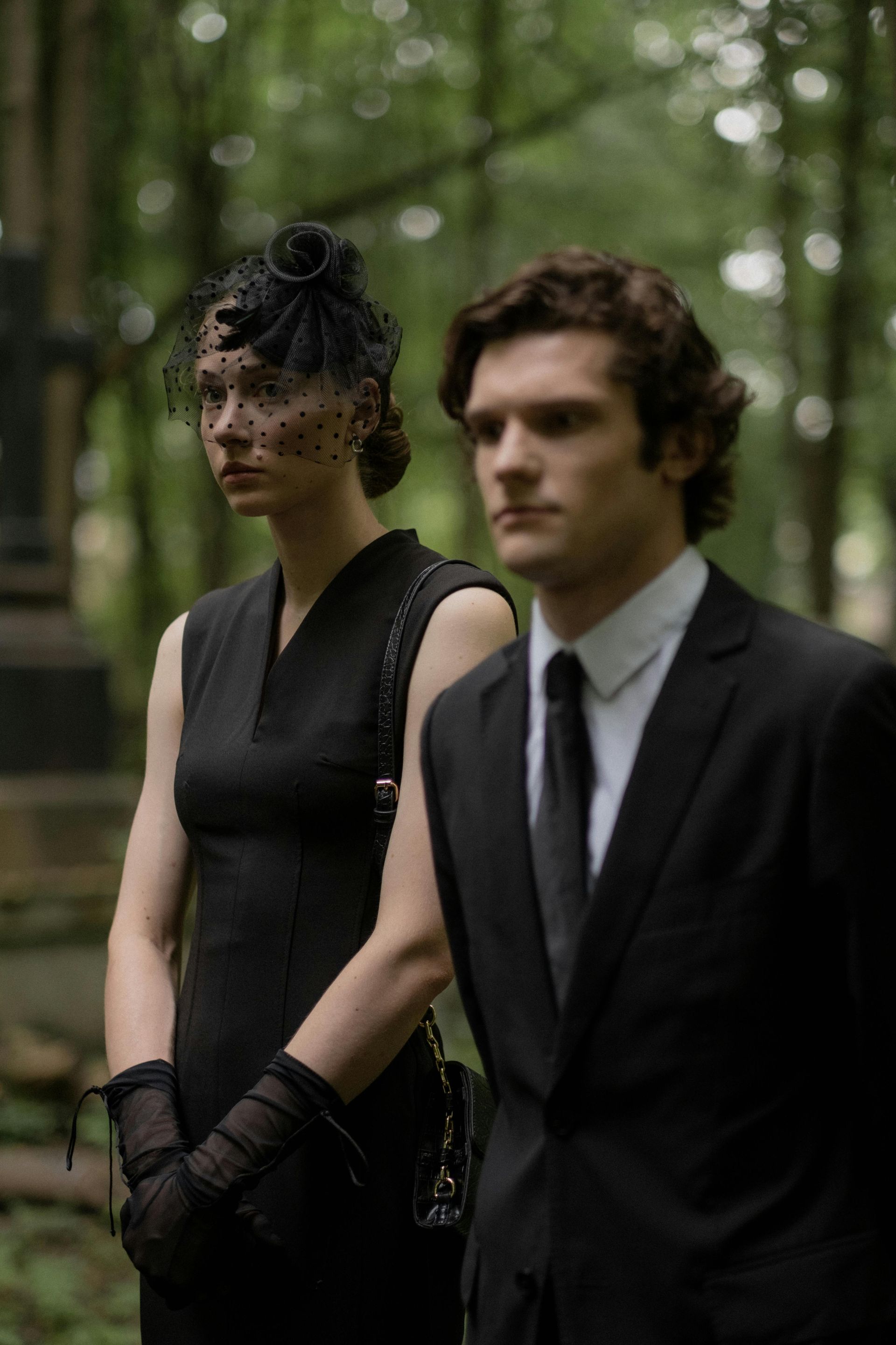 Two formally dressed people standing in a forest, one in black lace and one in a black suit.