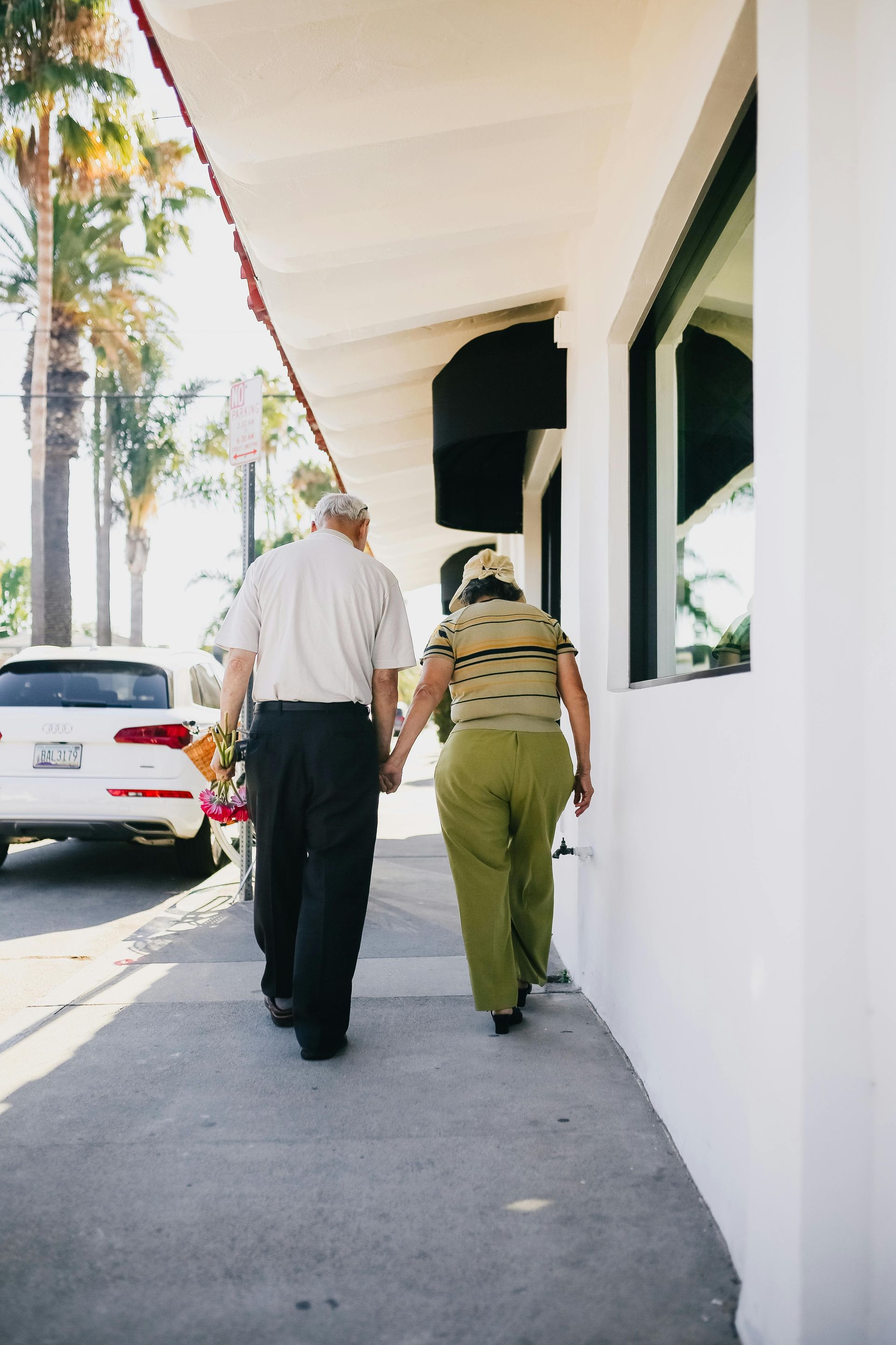 Two people walking along a sunny sidewalk beside white buildings and parked cars