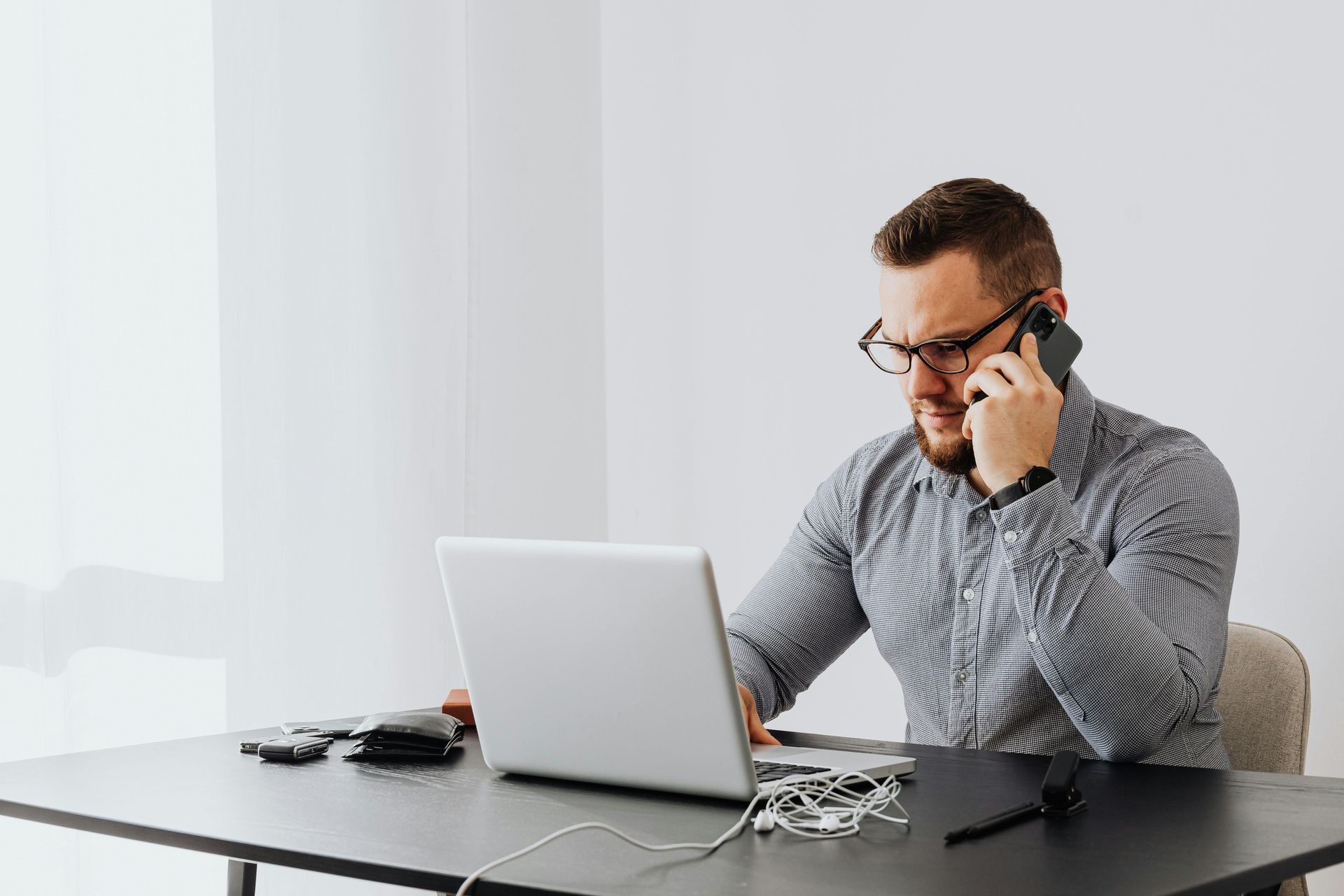 Man on a laptop talking on a cellphone at a desk in a bright office
