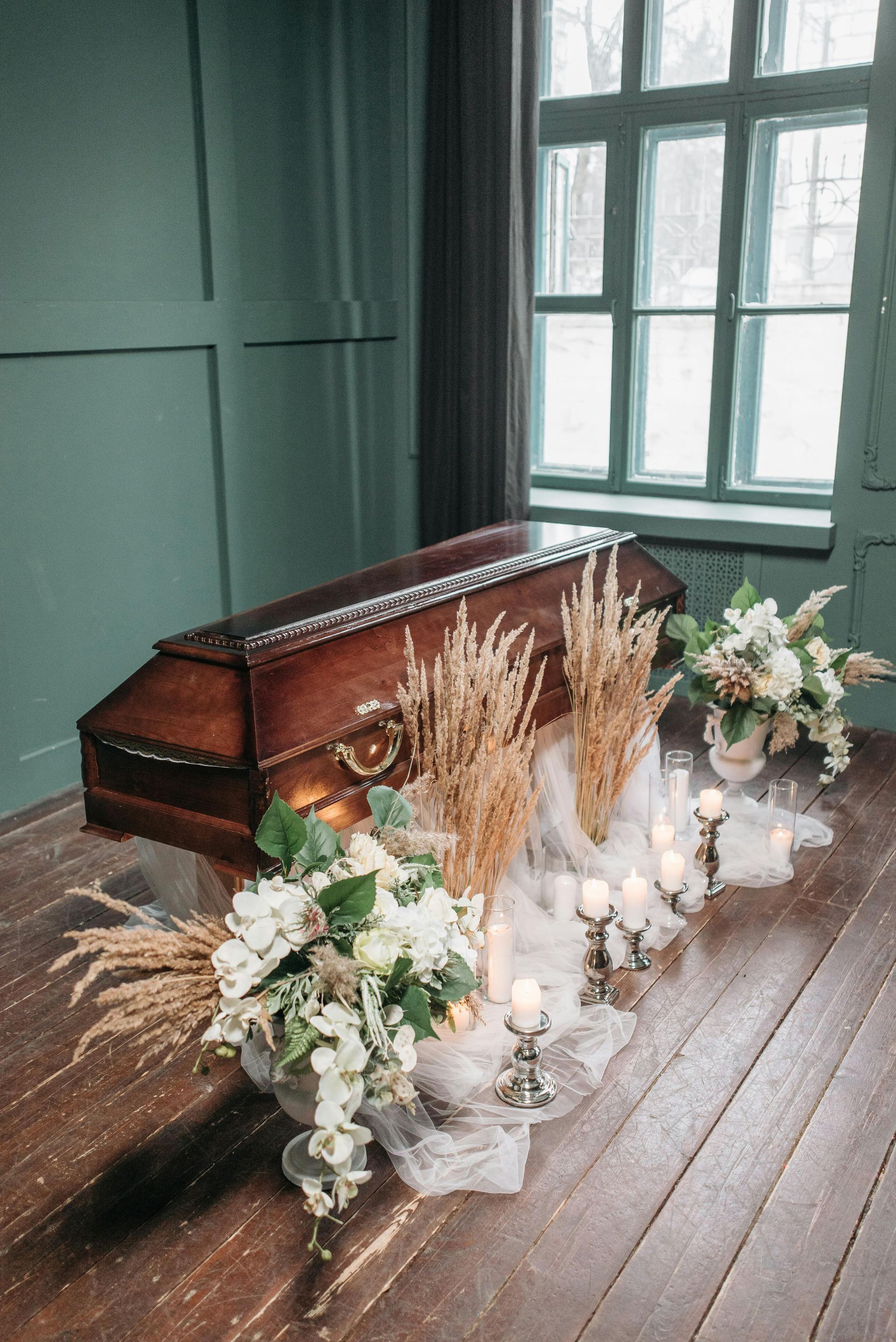Wooden coffin surrounded by white flowers, candles, and dried grasses in a dim green room