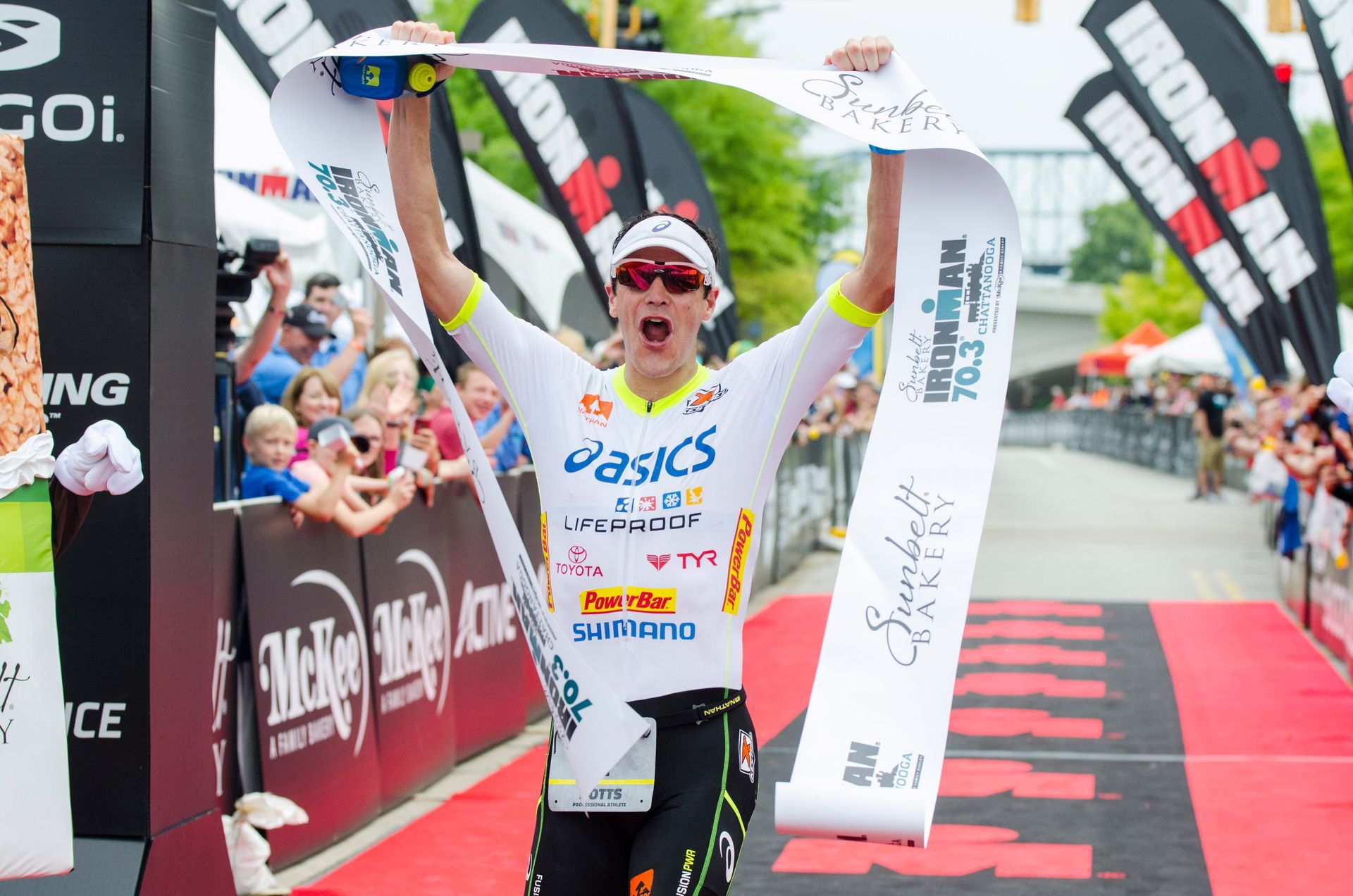 Runner in white crosses finish line with arms raised, winning a road race amid cheering crowd and banners.