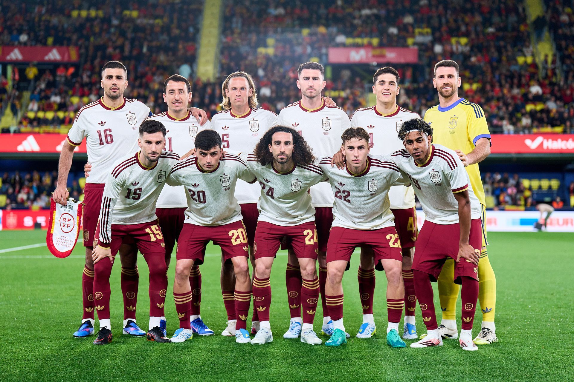 Football team posing for a group photo on the pitch, wearing white shirts and maroon shorts.