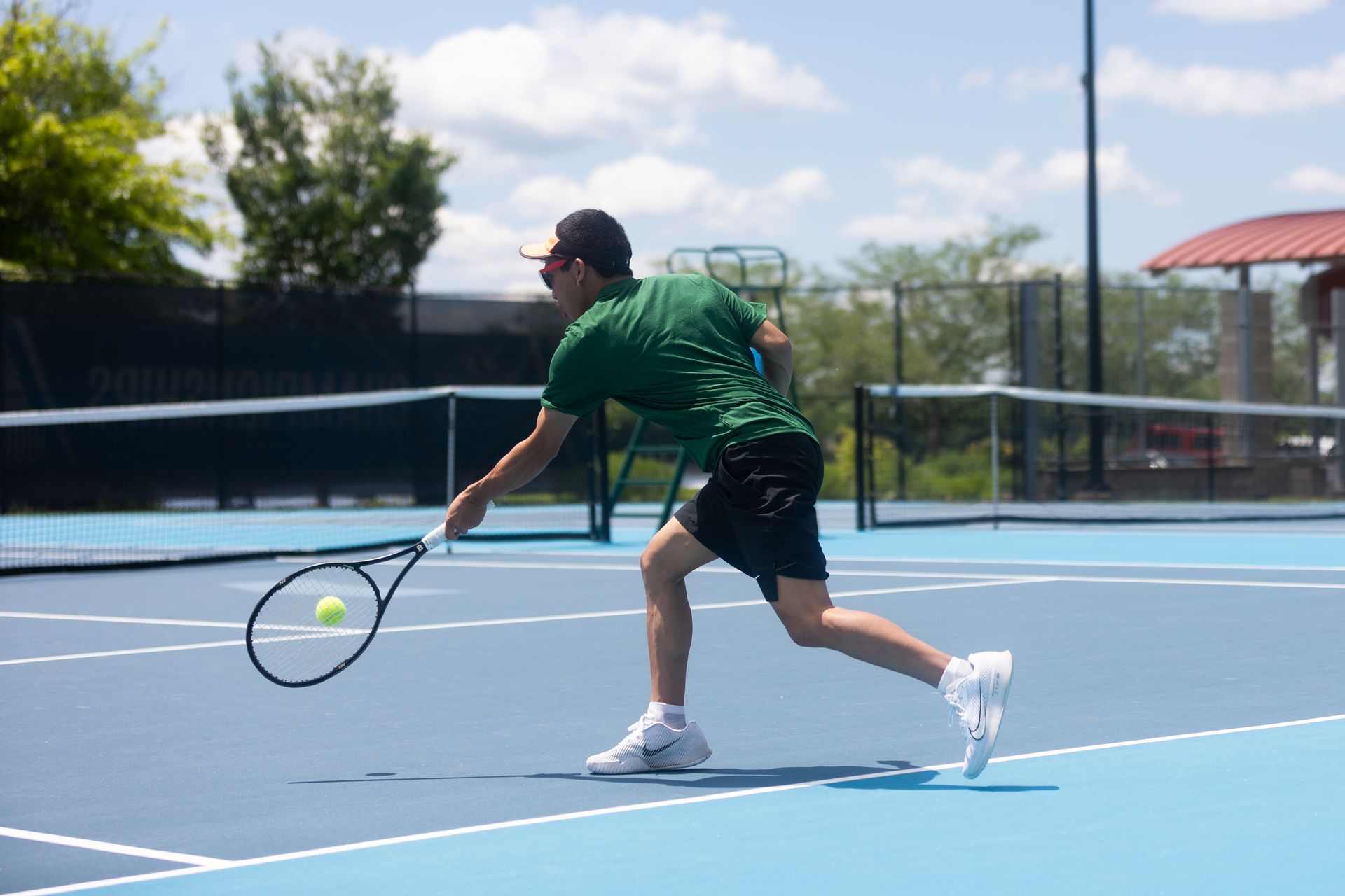Tennis player in green shirt lunging for a low ball on a blue court outdoors