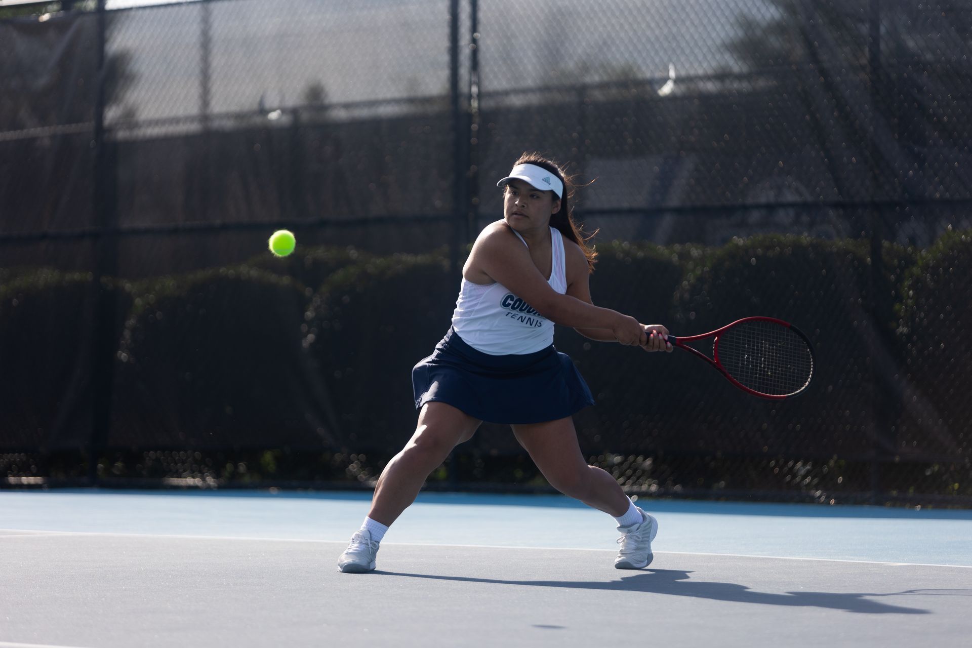 Tennis player in white and blue swings a racket on an outdoor court as the ball approaches.