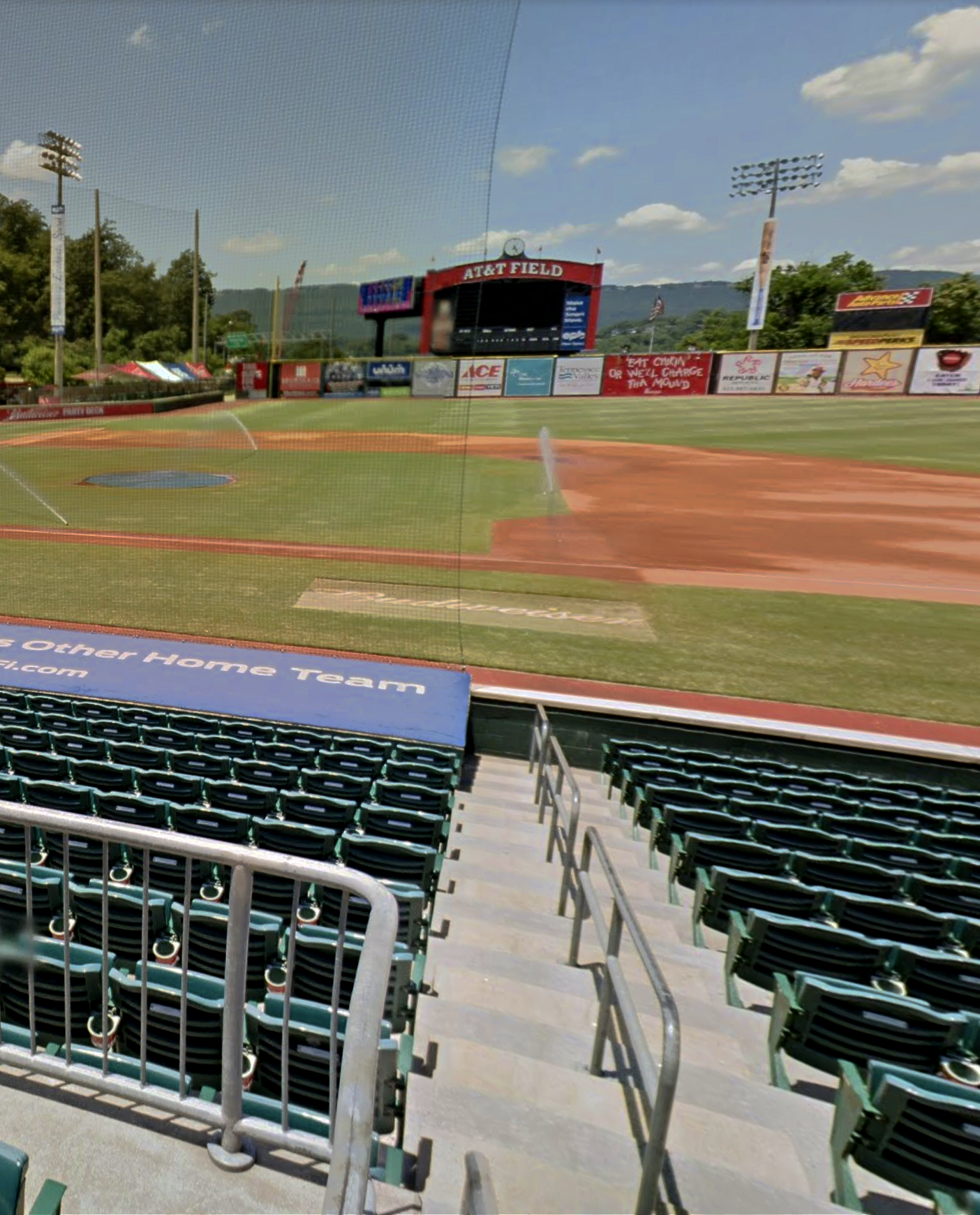 Empty baseball stadium with green seats, infield, scoreboard, and clear blue sky.