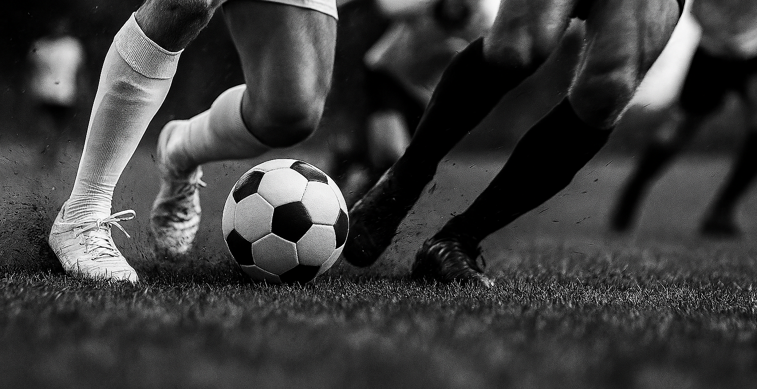 Soccer players’ legs in motion around a ball on a field, black-and-white close-up.