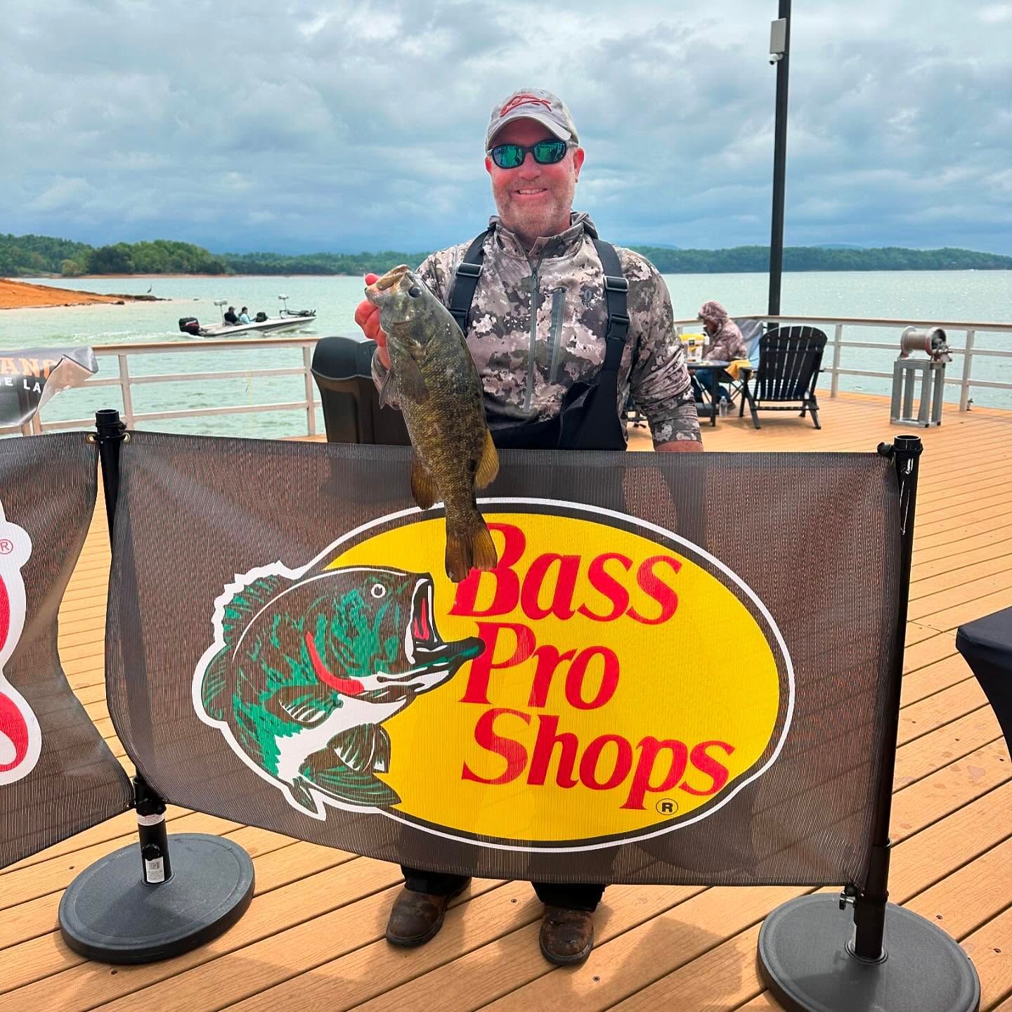 Man holding a large fish beside a Bass Pro Shops banner on a dock with water in the background