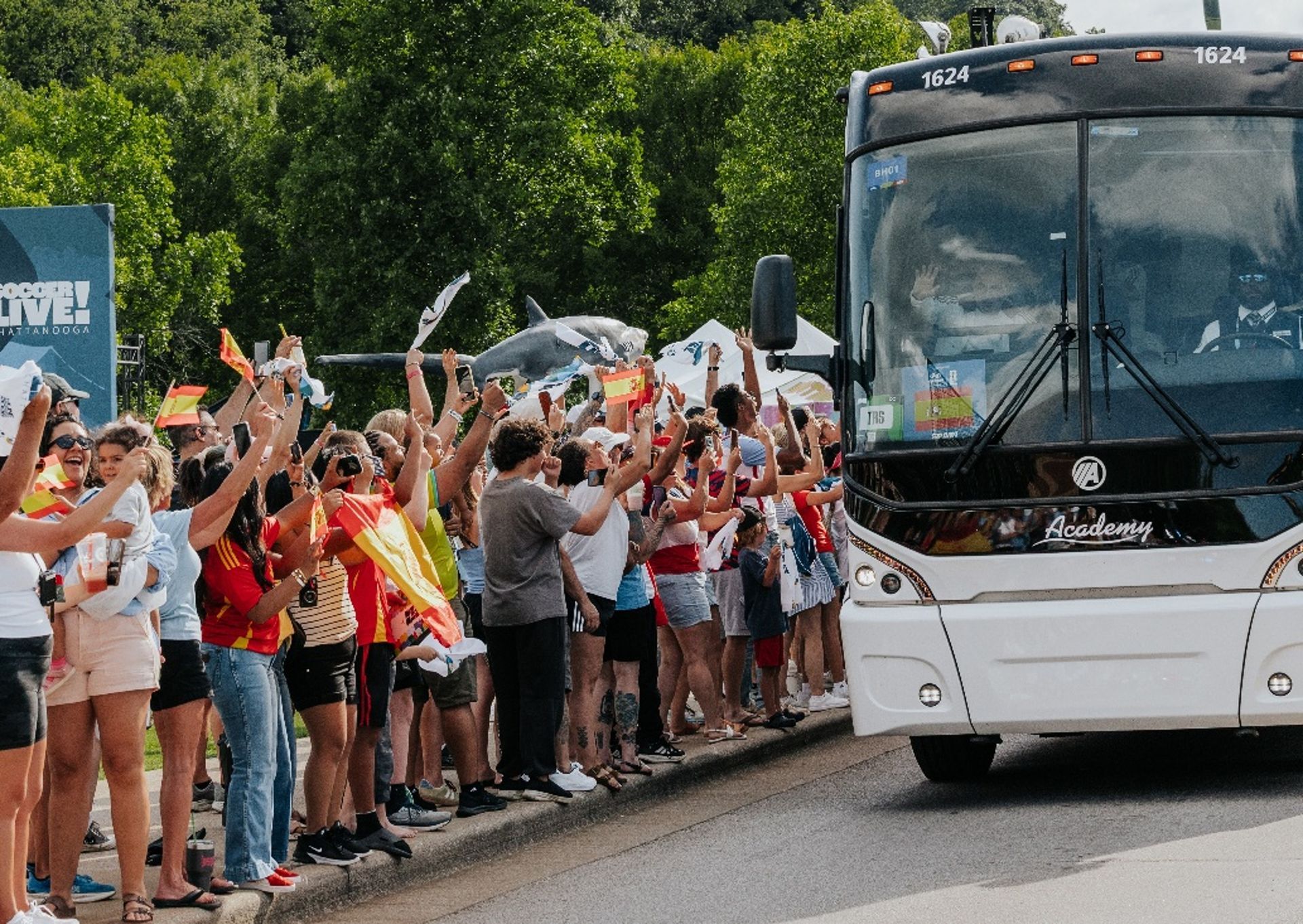 Crowd cheering and waving near a large bus on a road, with trees in the background