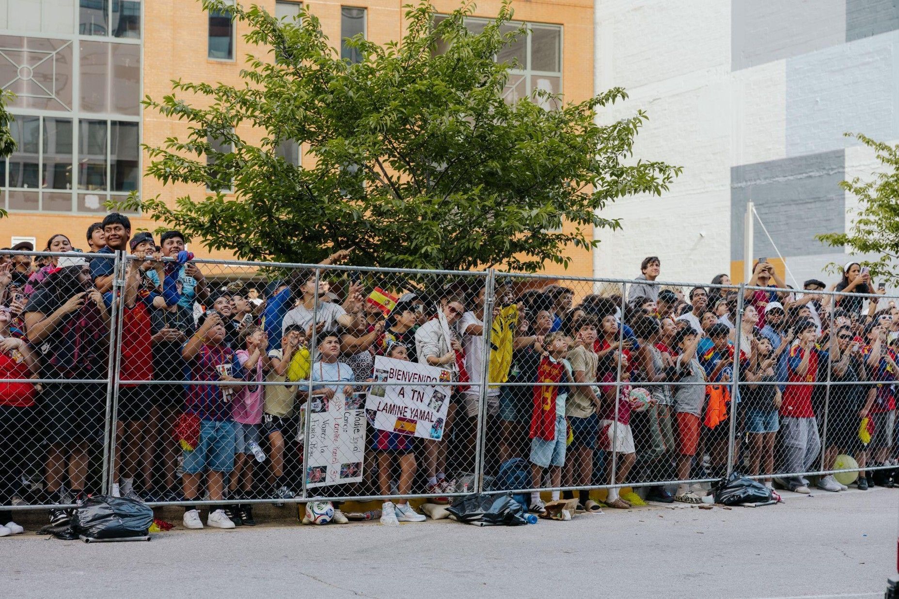 Crowd lined behind a chain-link fence at an outdoor event, holding signs in front of a building.