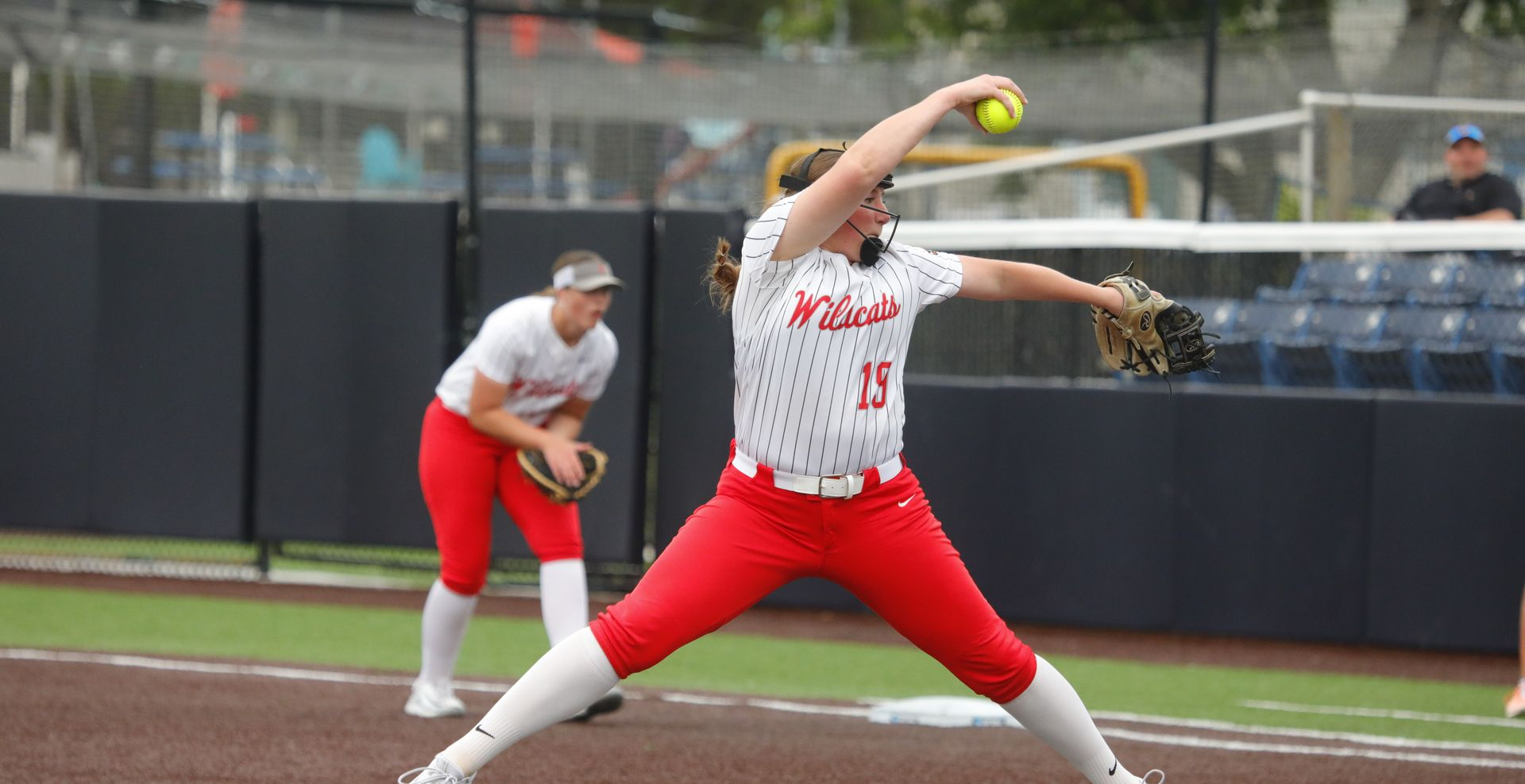 Softball pitcher in red and white uniform mid-throw on field, with catcher behind