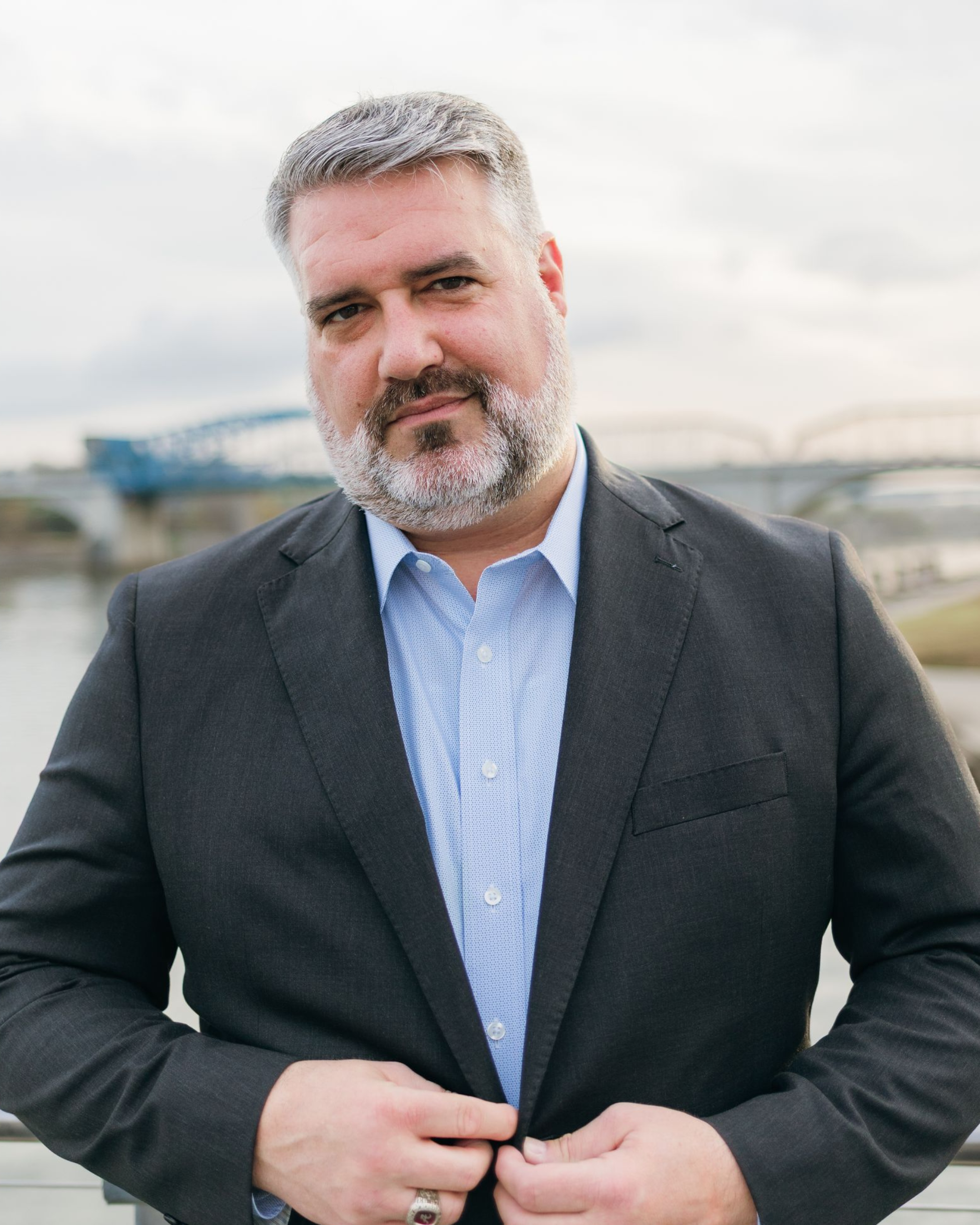 Man in a dark blazer and blue striped shirt buttoning his jacket outdoors with a blurred bridge behind him