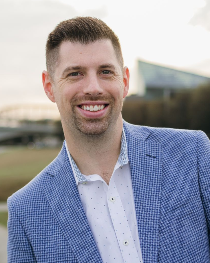 Smiling man in a blue blazer and white shirt standing outdoors with a blurred building behind him