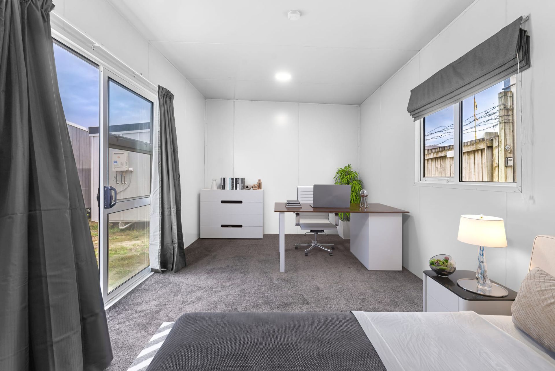 Bright minimalist bedroom with a bed, desk, chair, dresser, and large windows with gray curtains.