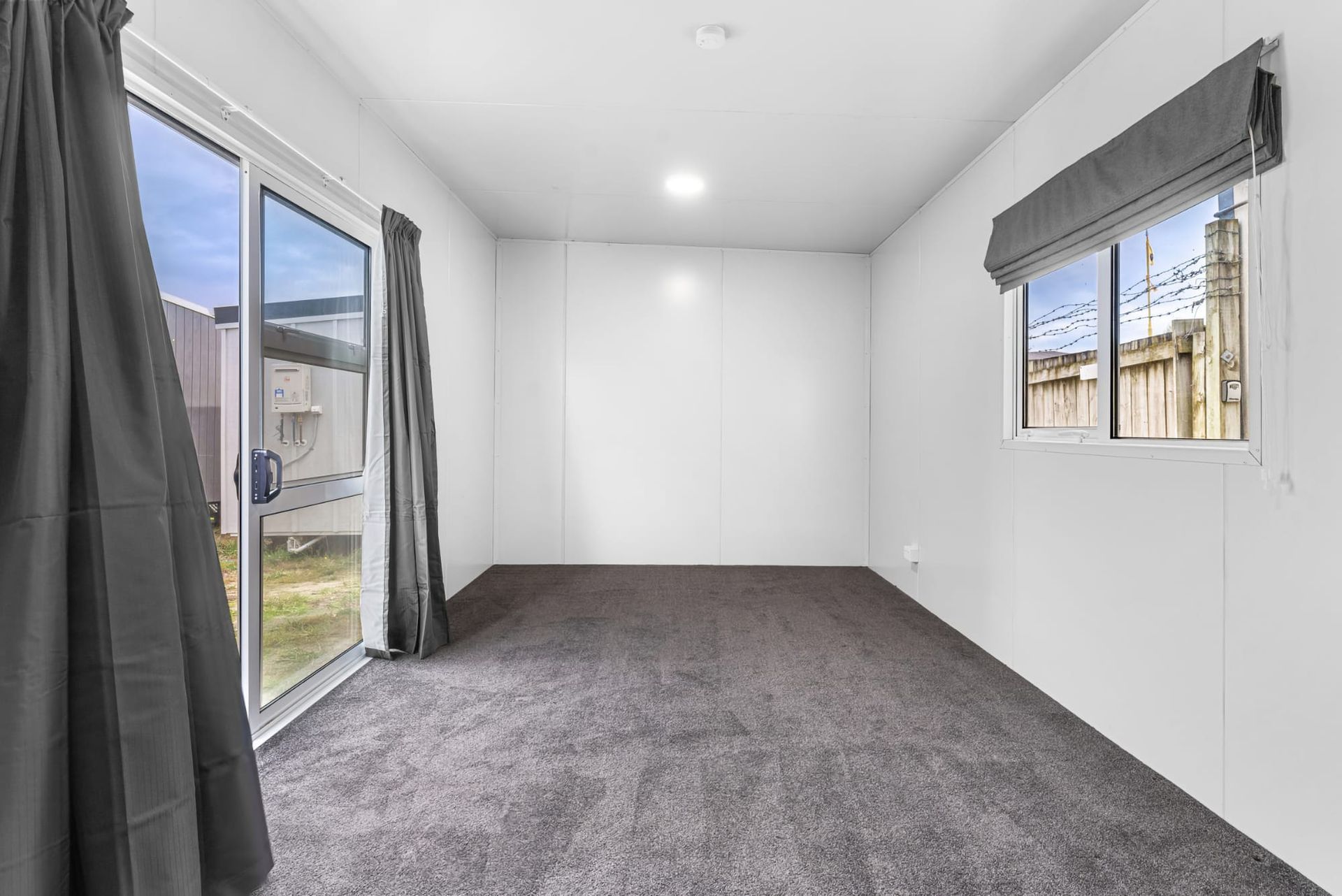 Empty carpeted room with white walls, open door to outside, and two windows with gray blinds