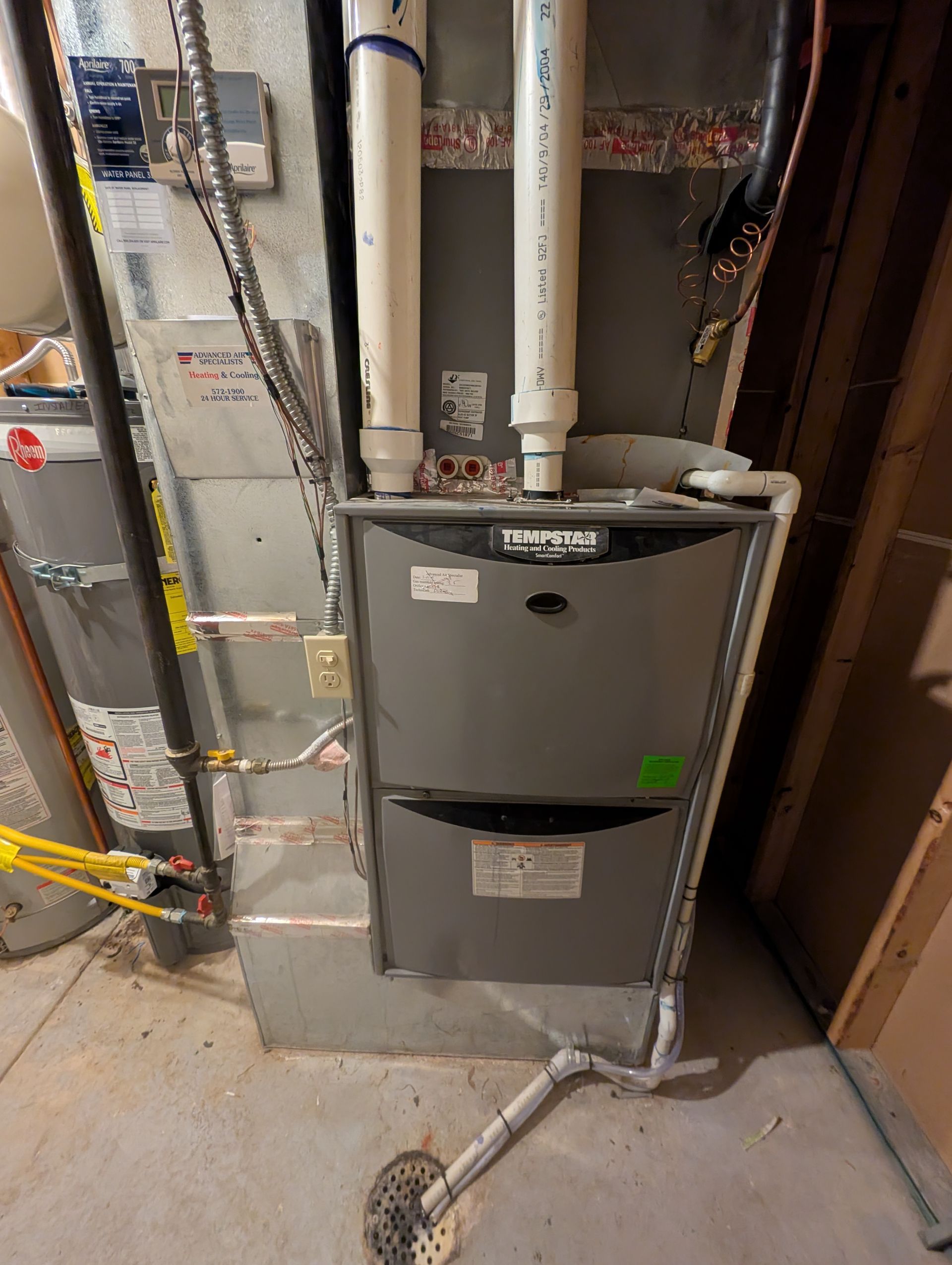 Gray furnace and ducts in a basement utility room with exposed pipes and wiring.