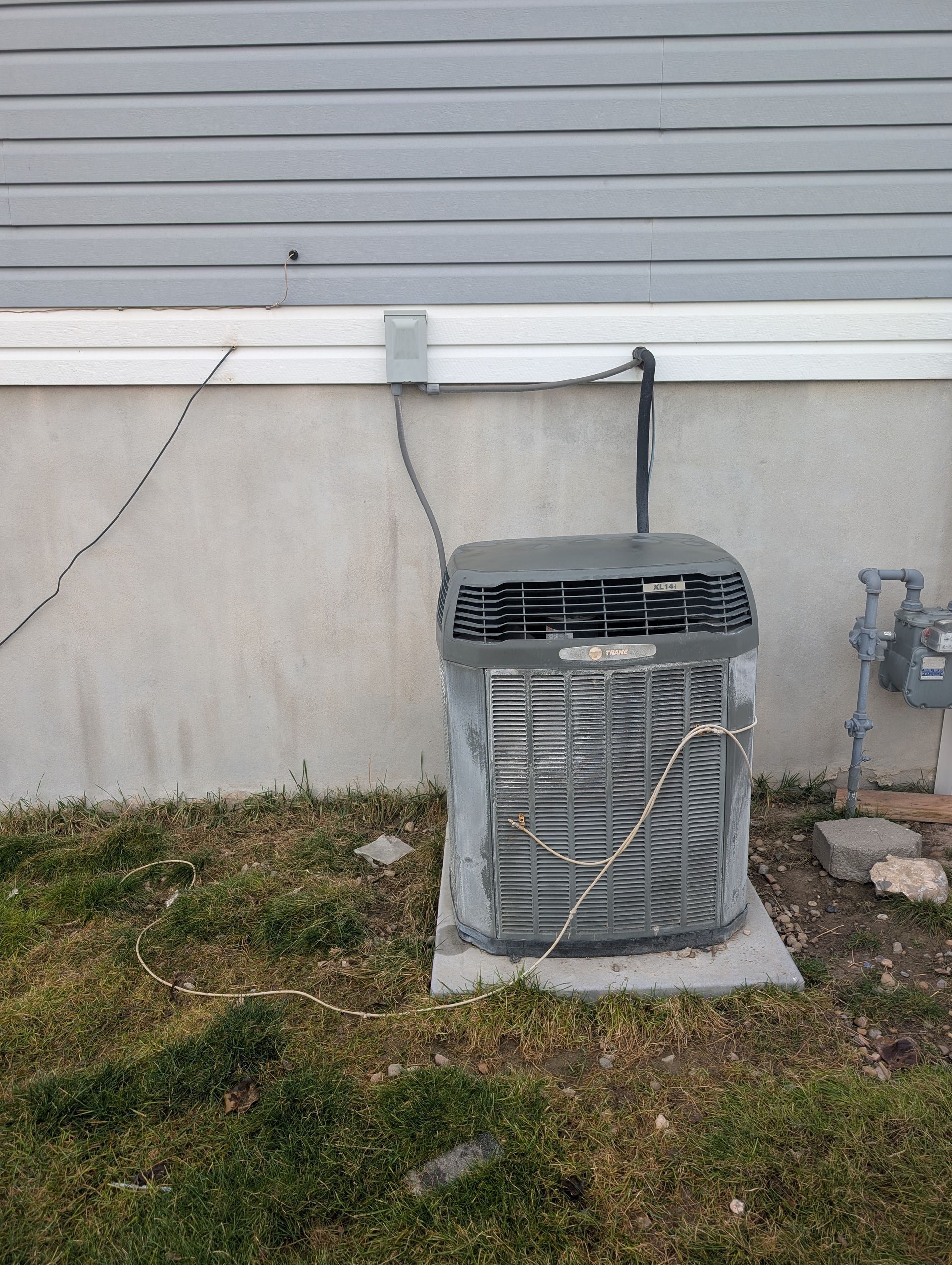 Outdoor HVAC condenser unit beside a gray house wall and utility line on a concrete pad