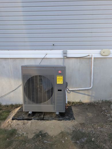 Outdoor heat pump unit beside a house wall, with gray siding and gravel ground.