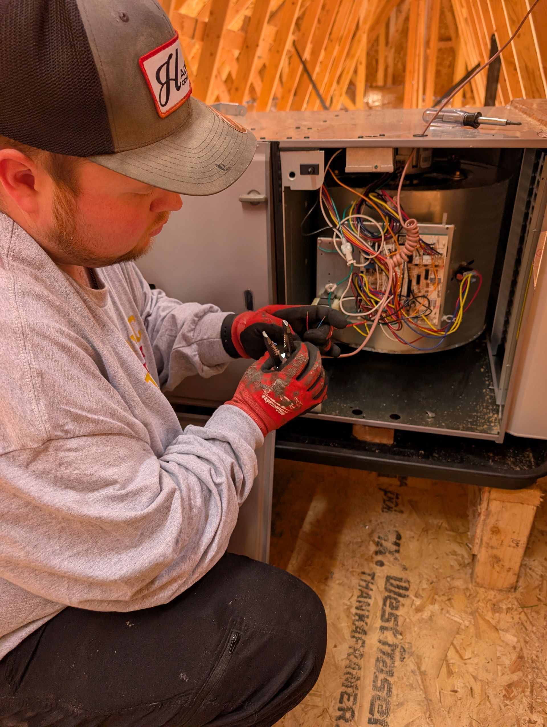 Technician in cap and gloves repairs wiring inside an open furnace cabinet in a wooden floor attic.