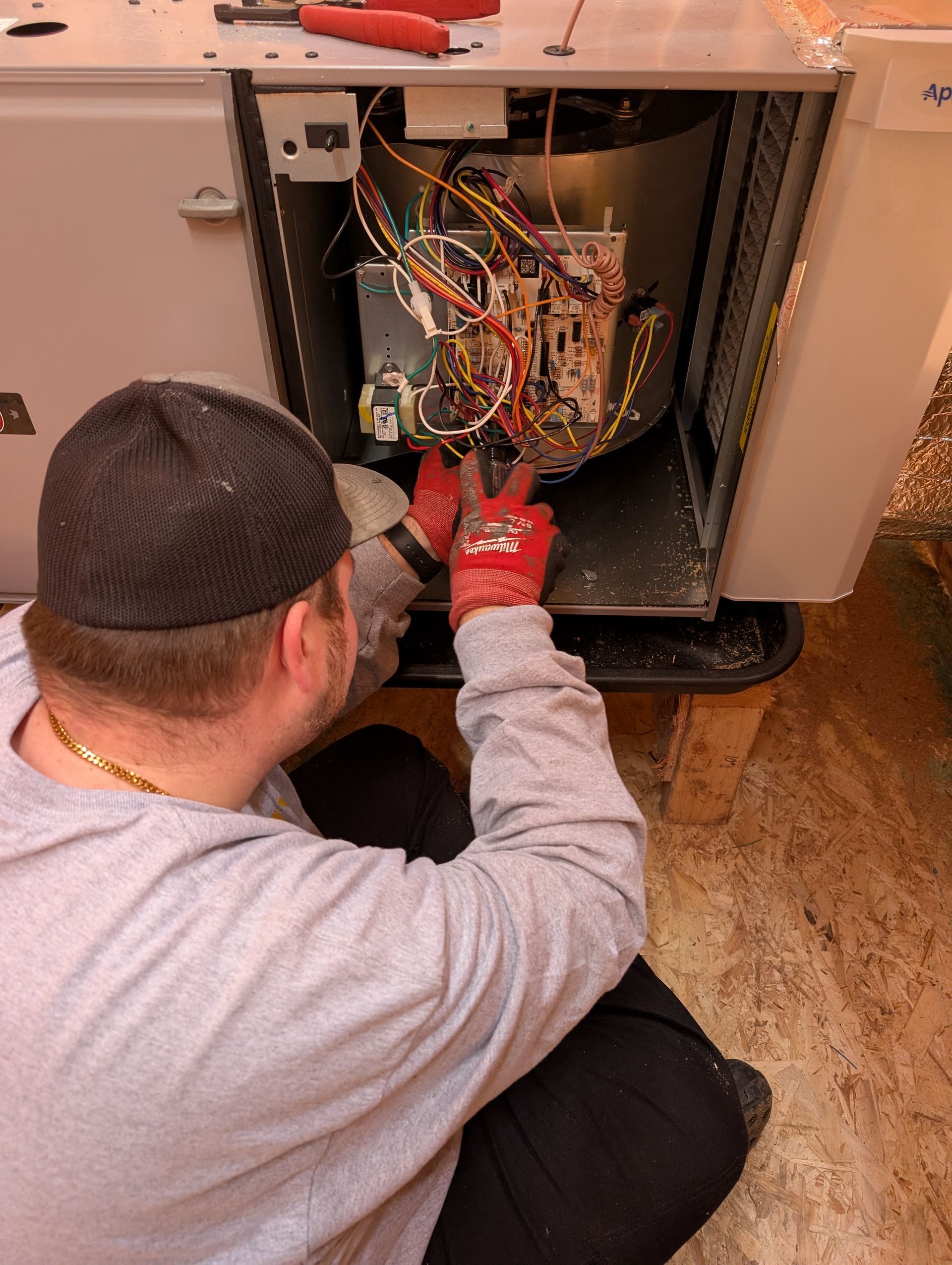 Technician kneels beside an open appliance, using a red tool to inspect exposed wiring.