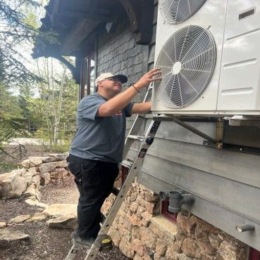 Technician on a ladder inspecting an outdoor air conditioning unit beside a house.