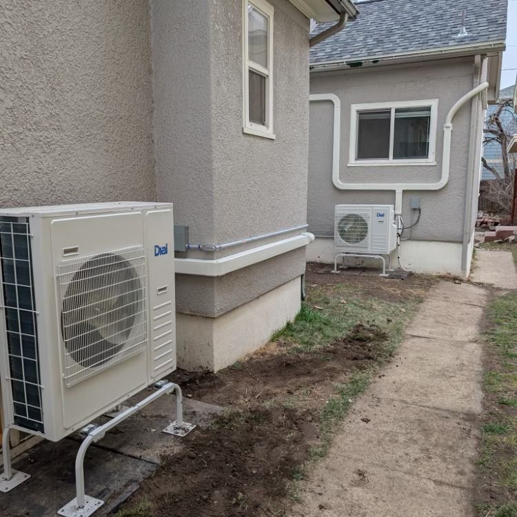 Two outdoor HVAC units beside a house along a narrow dirt path in a small yard