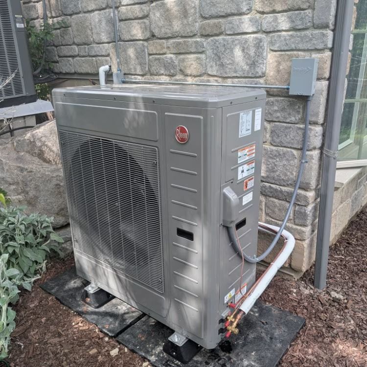 Outdoor gray HVAC condenser unit beside a stone house wall, with pipes and wiring attached