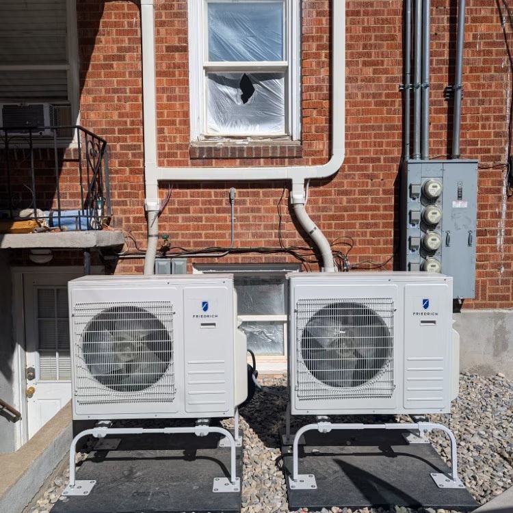 Two white outdoor AC units beside a brick house, with a broken window above and utility meters on the right.