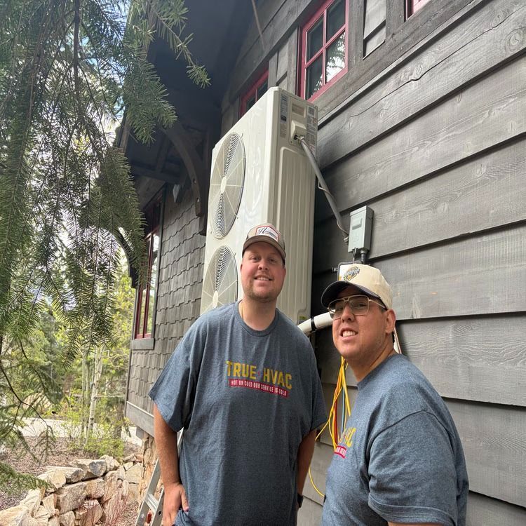 Two workers stand beside a wall-mounted HVAC unit on the side of a wooden house.