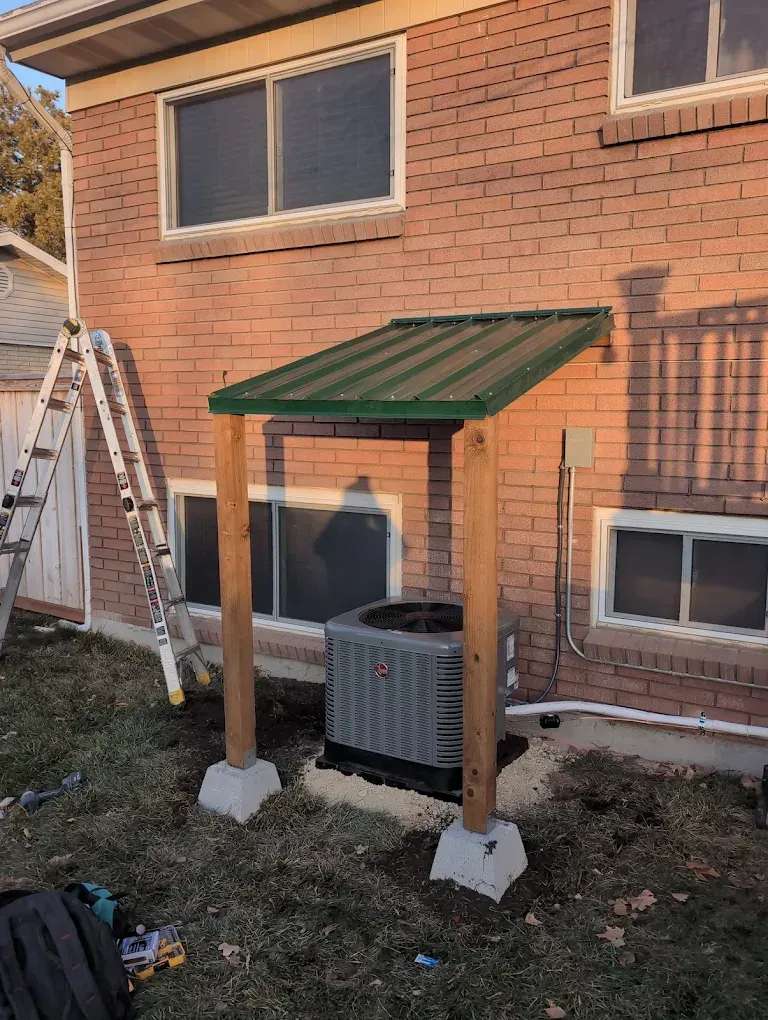 Backyard brick house with a small green-roofed shelter over an air conditioner, ladder nearby