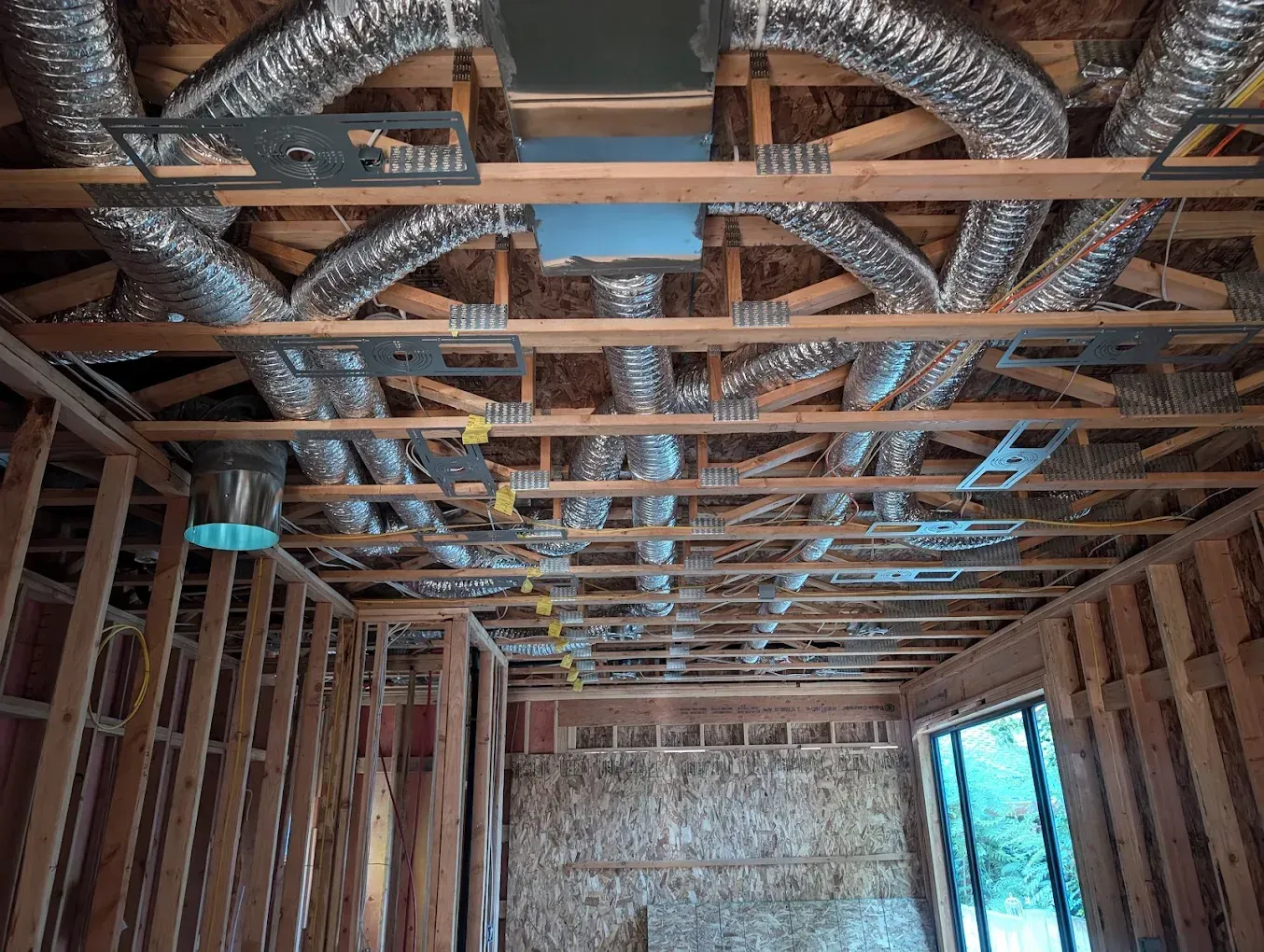 Exposed ceiling framing with ductwork and wiring in an unfinished room, with a window on the right.
