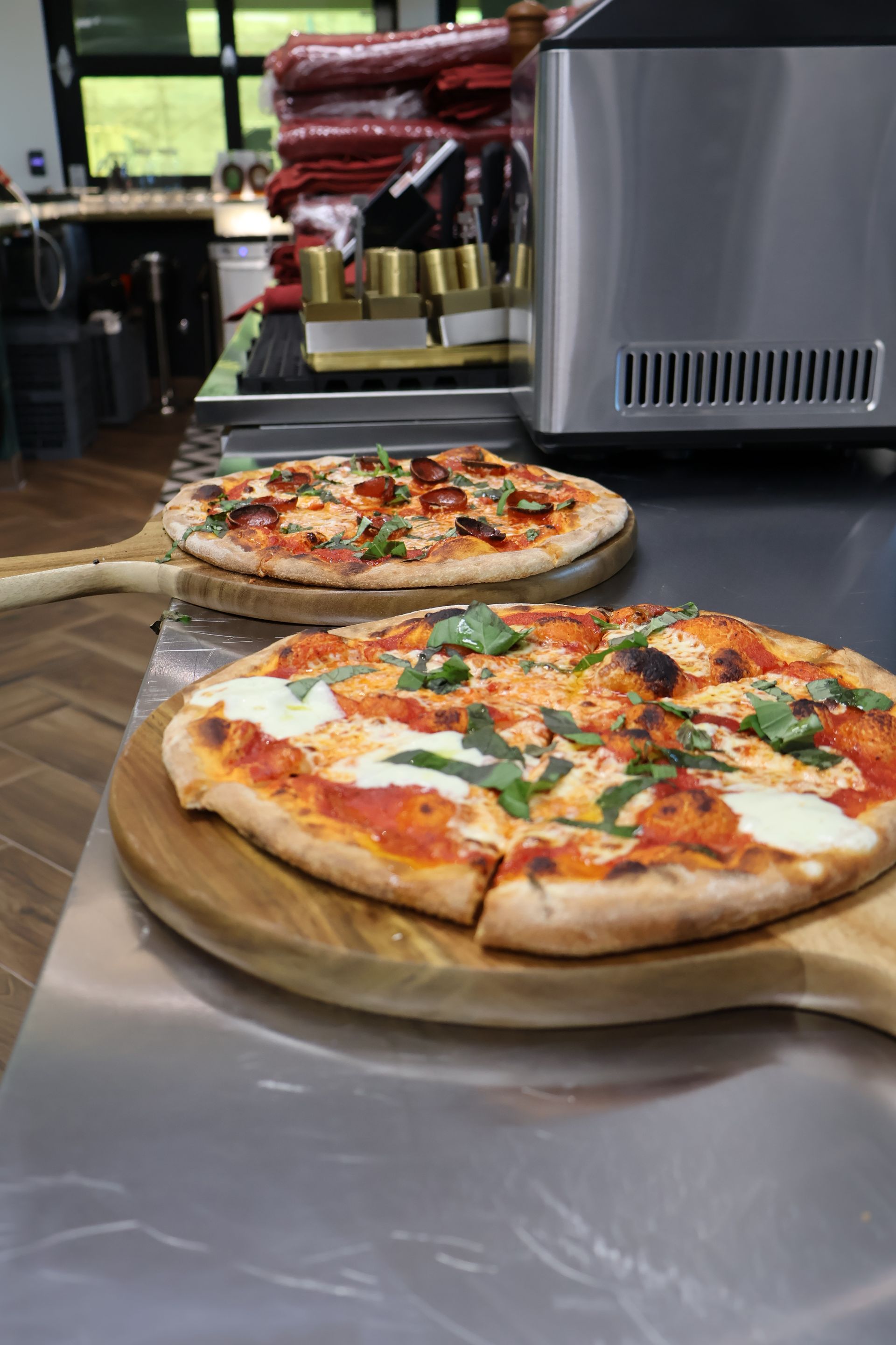 Two pizzas on wooden serving boards rest on a stainless steel counter in a restaurant kitchen.